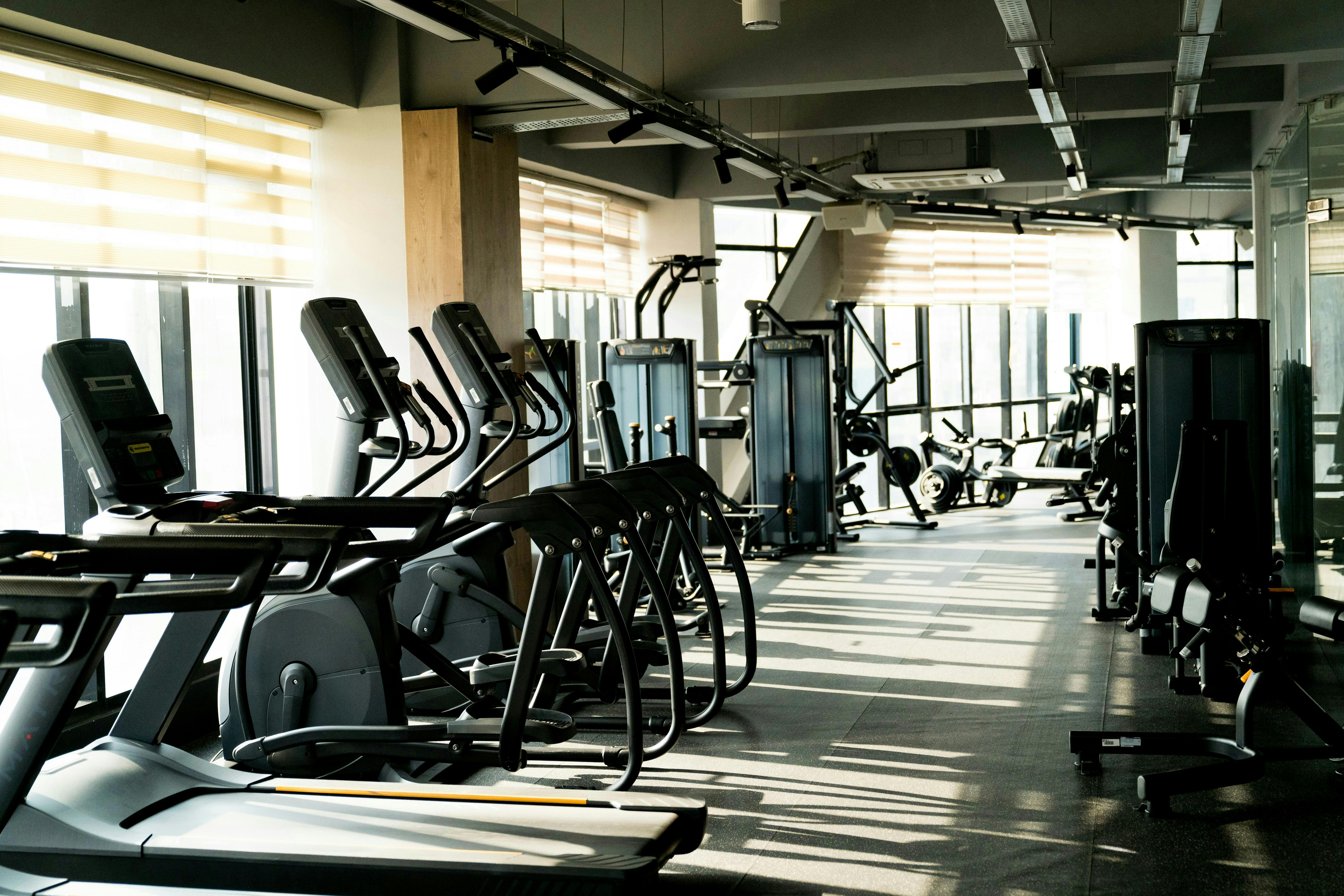 A bright, sunlit gym interior featuring a row of elliptical machines and treadmills positioned in front of large floor-to-ceiling windows with horizontal blinds. Long shadows stretch across the floor from the morning or afternoon sun.