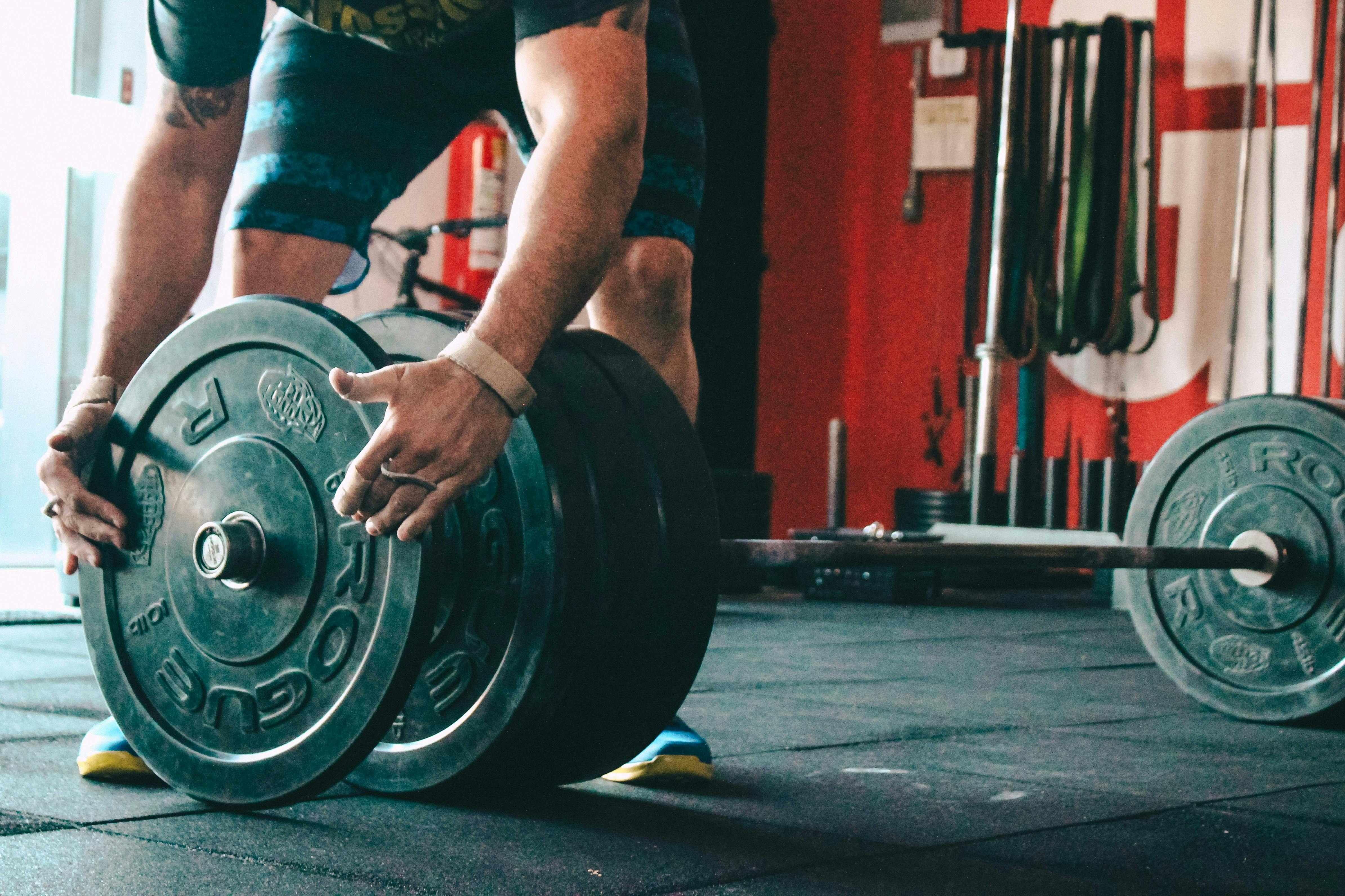 A close-up, low-angle shot of a person’s hands and forearms as they slide a heavy black Rogue weight plate onto a barbell. The background shows a gym wall with red accents and various resistance bands hanging.