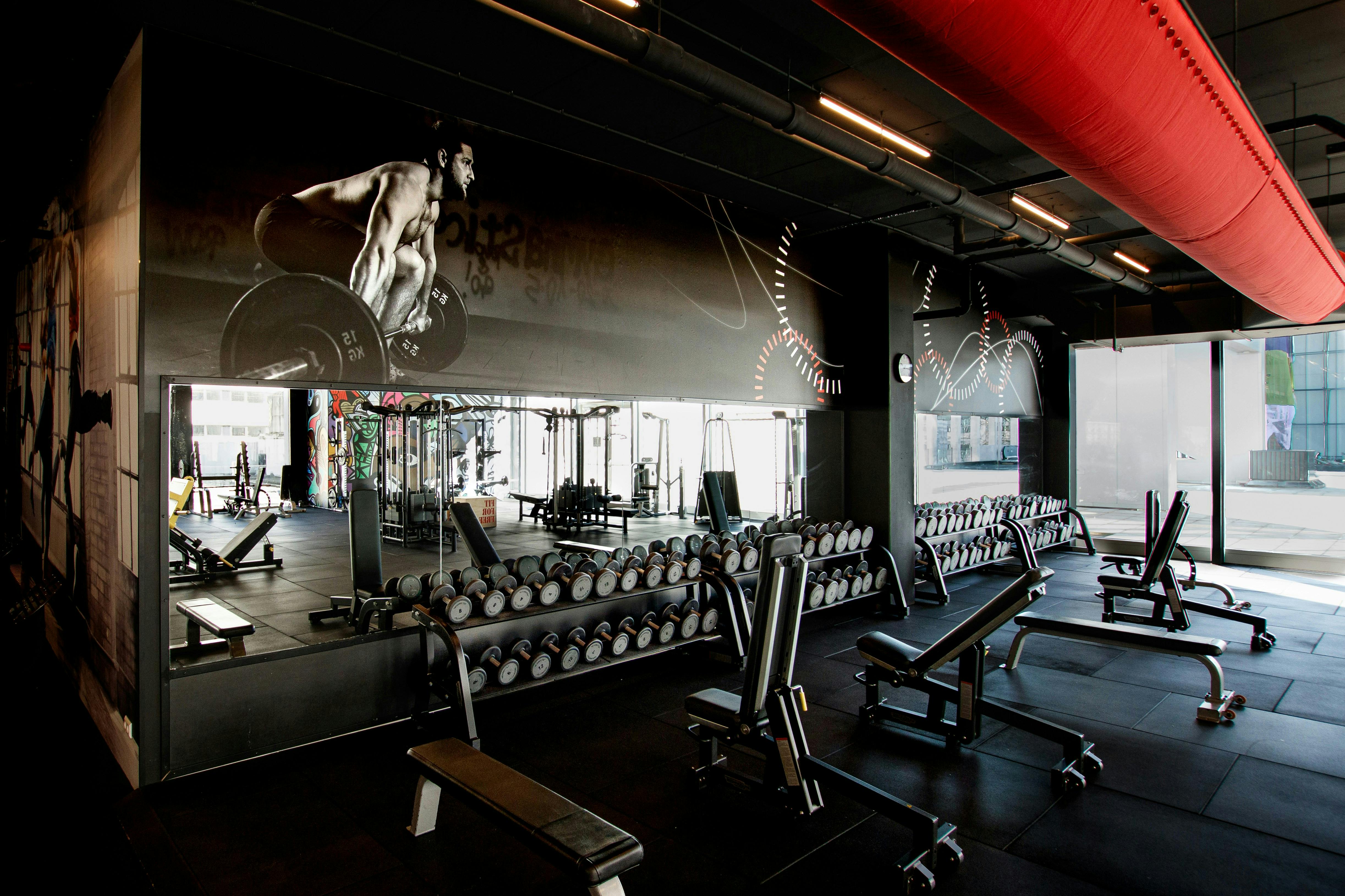 A spacious weight room featuring multiple rows of dumbbells on racks and several adjustable workout benches. A large black-and-white mural of a man lifting a barbell spans the back wall above a wide mirror.