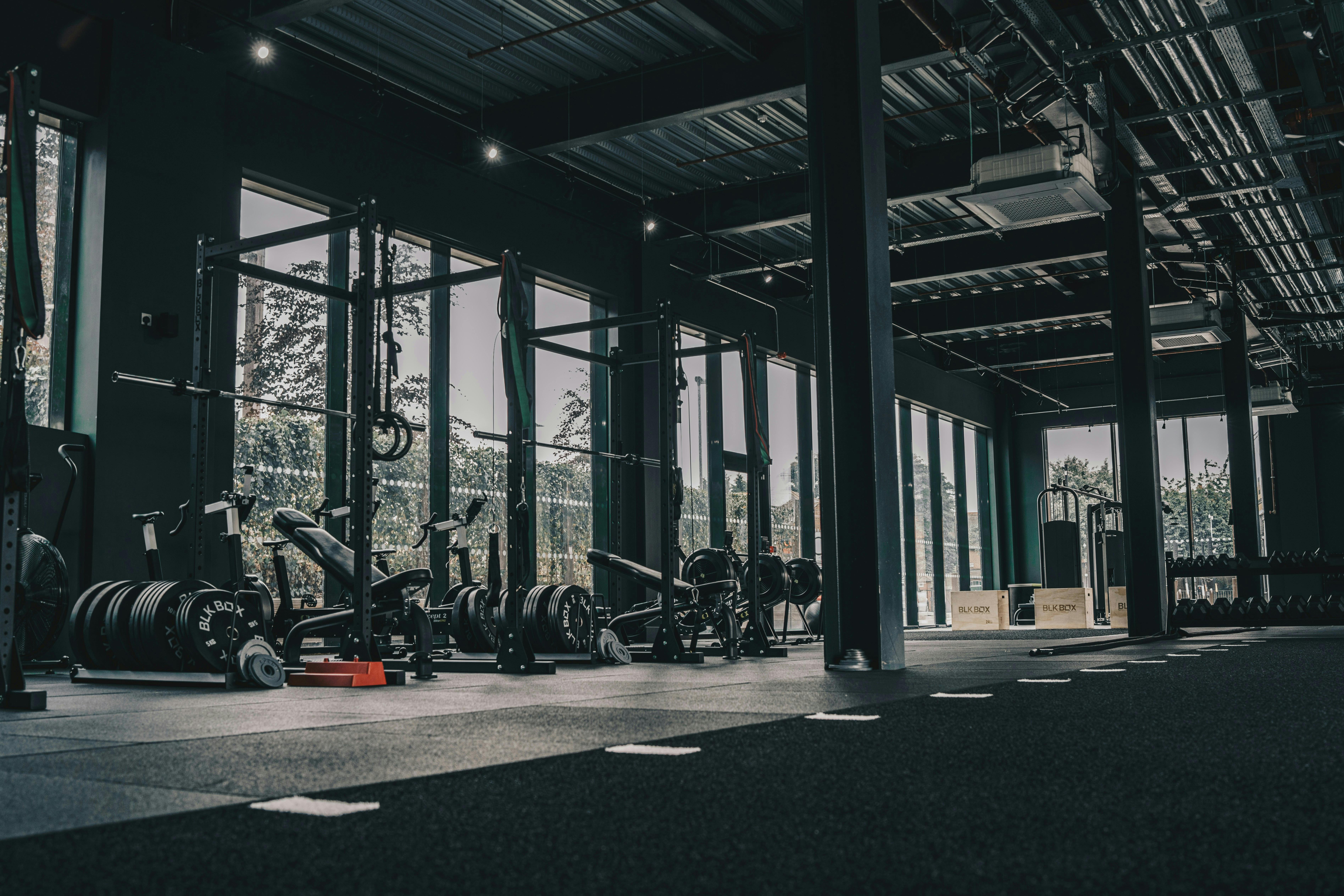 A moody, industrial-style gym with high ceilings and exposed pipes. Power racks and weight plates are neatly arranged on a black matted floor, with soft natural light coming through large windows on the left.