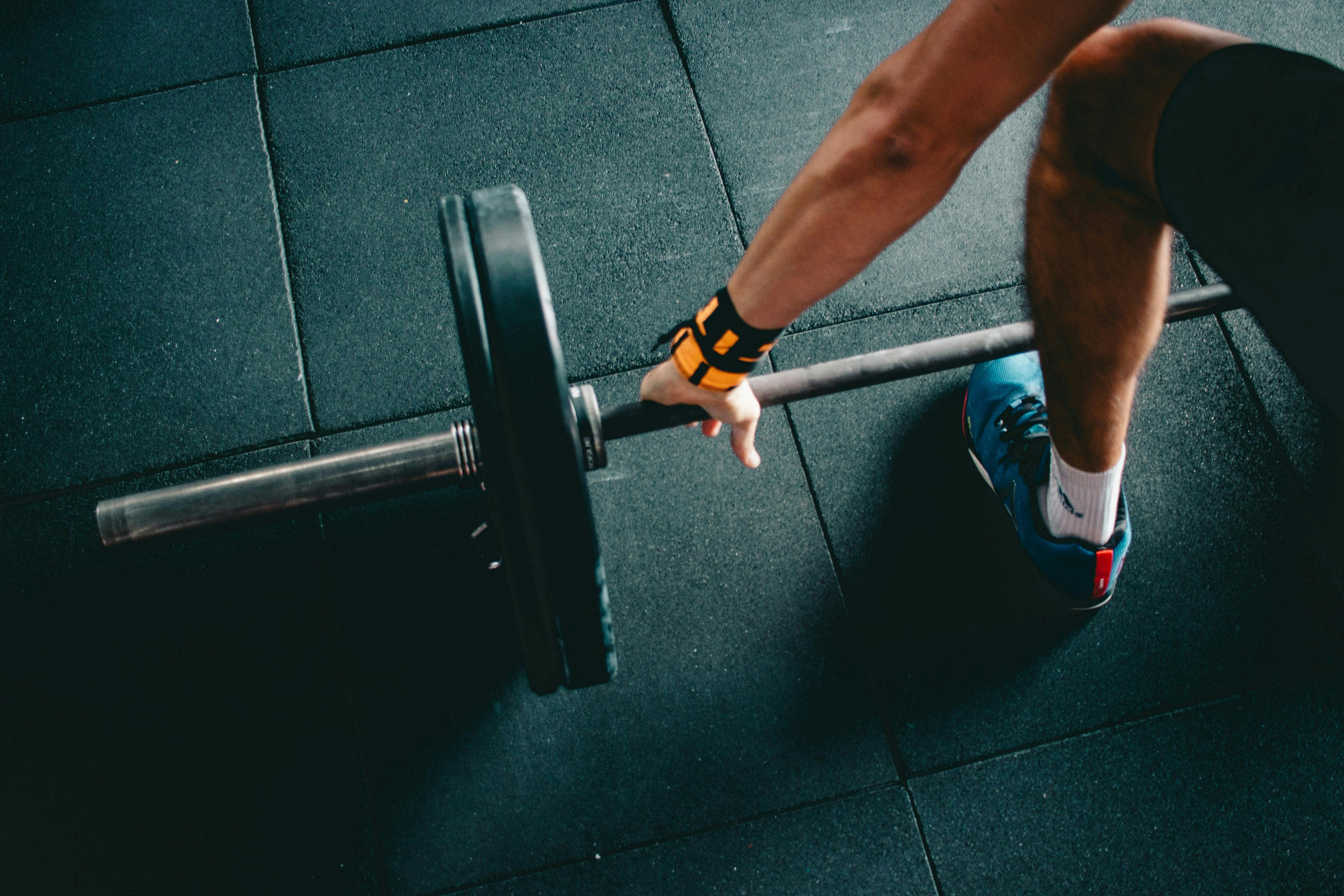 An overhead, high-angle view of an athlete's arm and leg as they prepare to lift a loaded barbell from a black rubber gym floor. The athlete is wearing a blue sneaker and a yellow wrist wrap.

