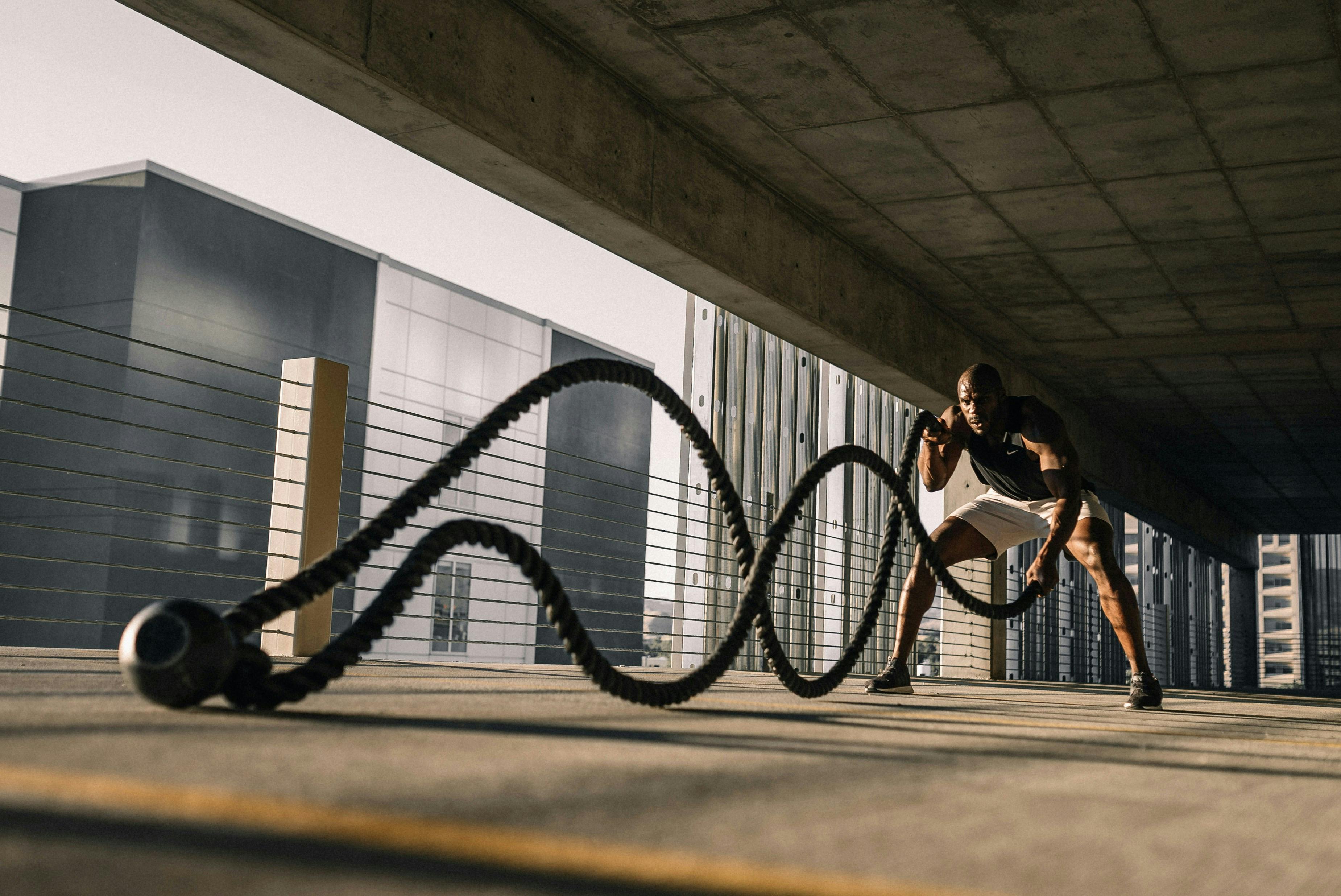 An outdoor, shaded workout area where a muscular man is mid-action using battle ropes. The ropes create a dynamic "S" wave pattern on the concrete floor, with a modern building and clear sky in the background.