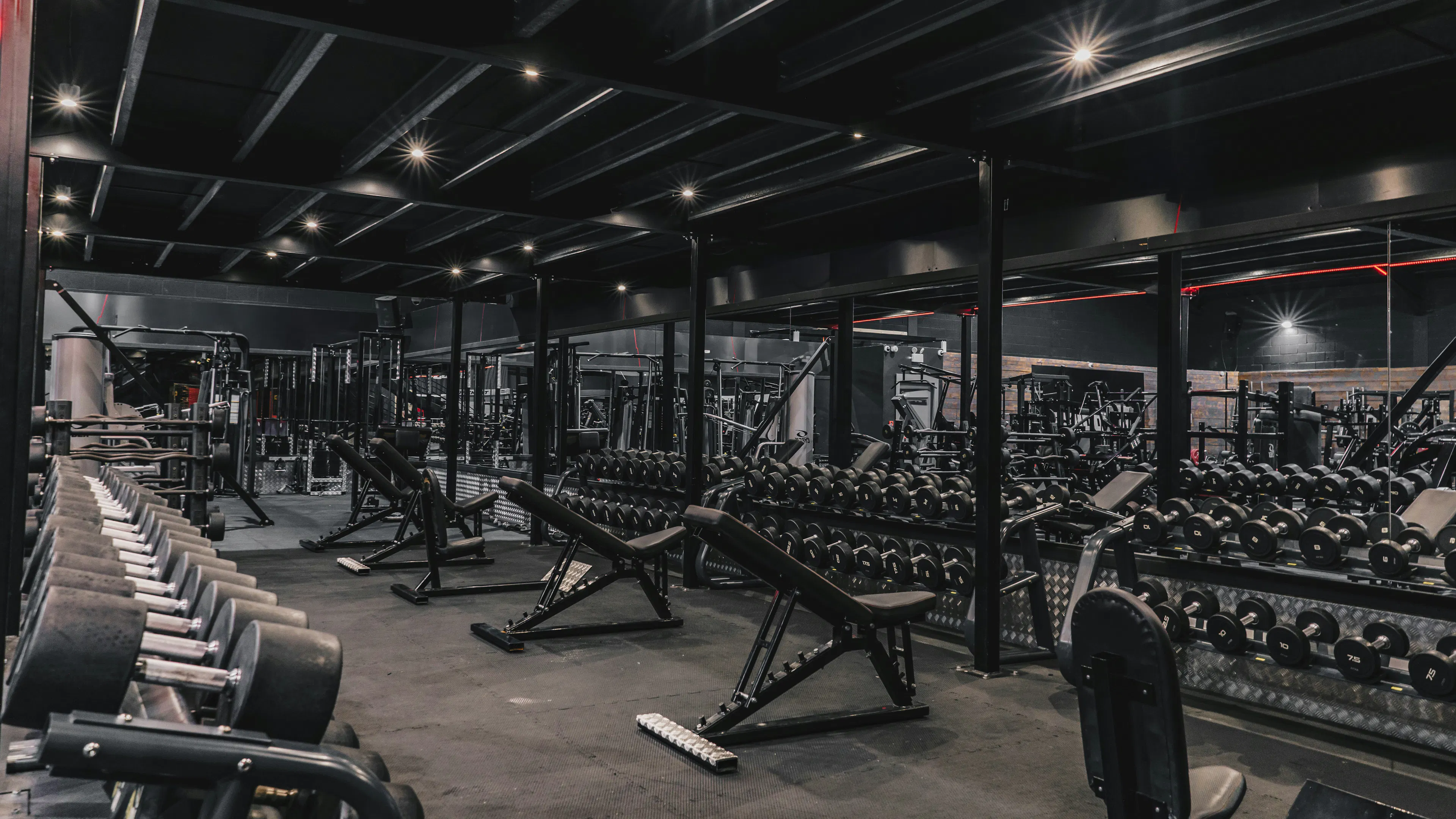 A professional gym setting featuring long racks of dumbbells and several incline weight benches. The room has a dark, sleek aesthetic with industrial black ceilings and spotlighting that reflects off the equipment and mirrors.
