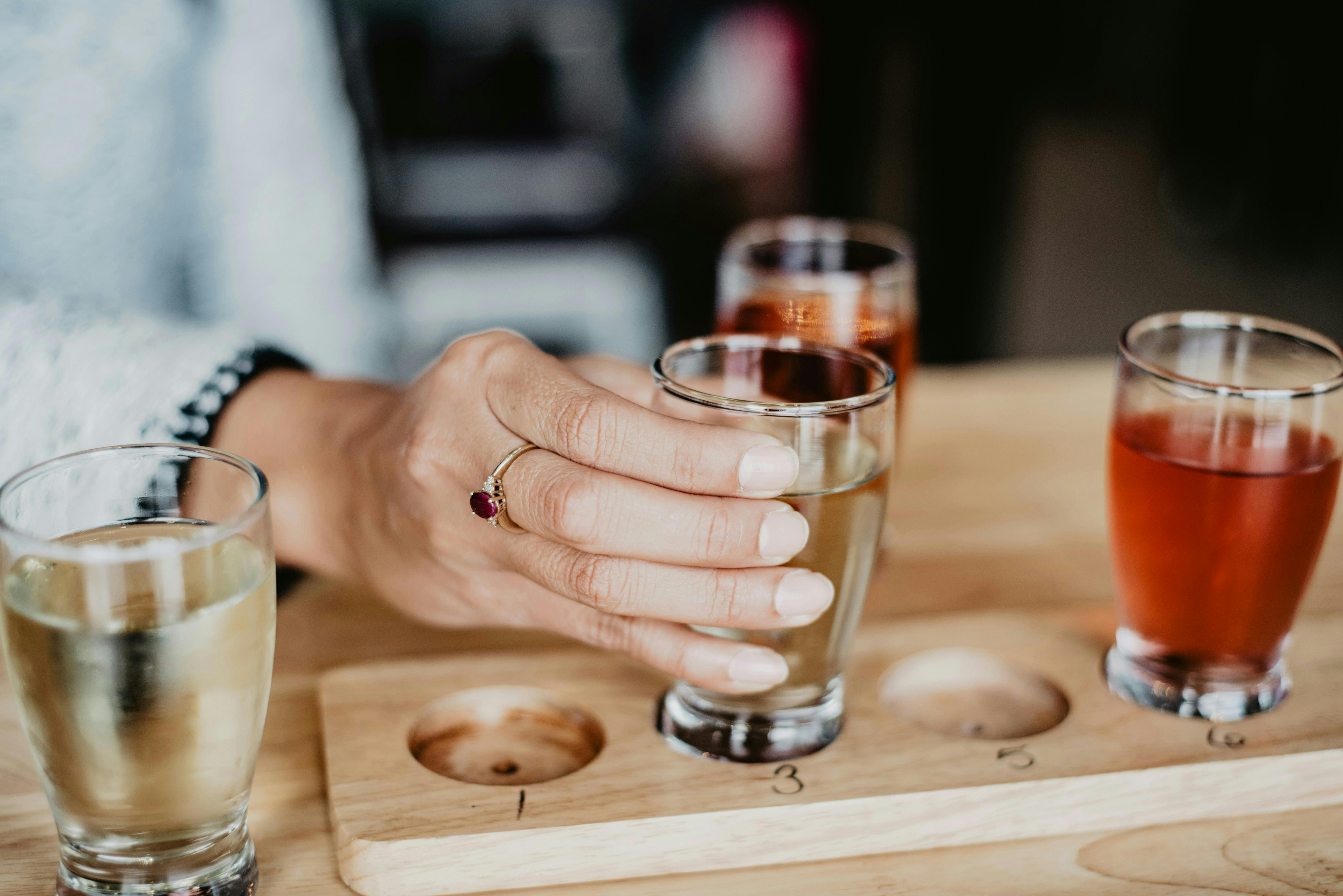 A close-up shot of a person's hand, wearing a gold ring with a red gemstone, reaching for a small glass of light-colored beer from a wooden flight paddle. Two other glasses filled with amber and reddish beer sit nearby.