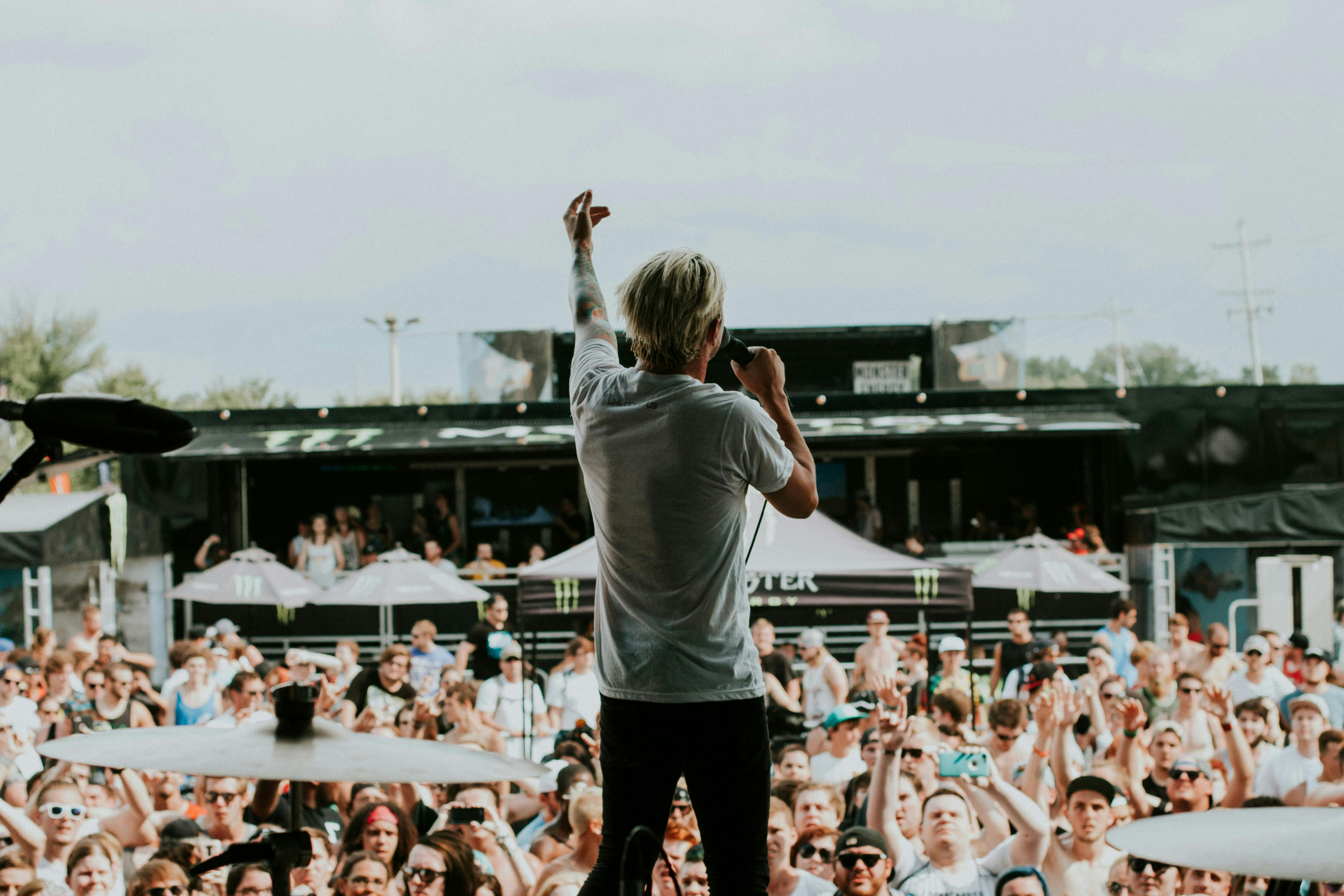 A view from behind a singer on stage performing for a large, sunlit outdoor crowd at a music festival. The singer has blonde hair and a white t-shirt, gesturing with one hand toward the audience.