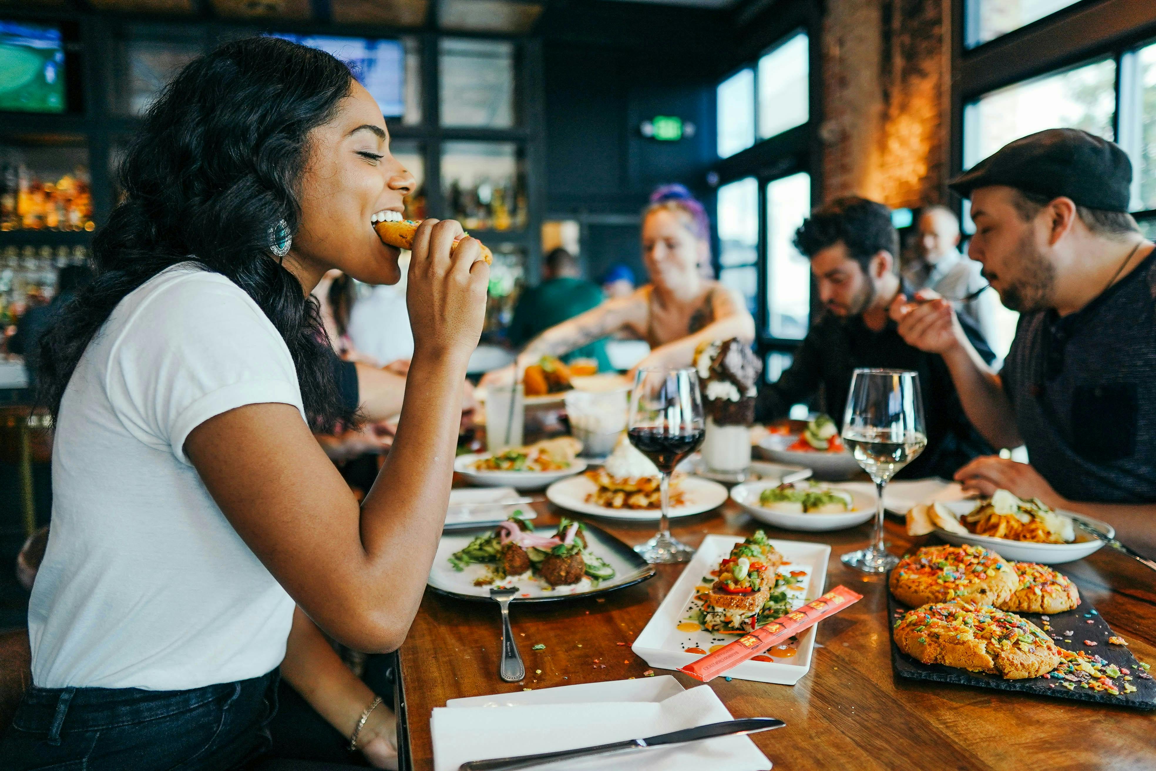 A group of friends enjoying a diverse spread of colorful dishes and drinks at a wooden restaurant table. In the foreground, a woman laughs while taking a bite of food, with plates of sliders, salads, and wine glasses blurred in the background.