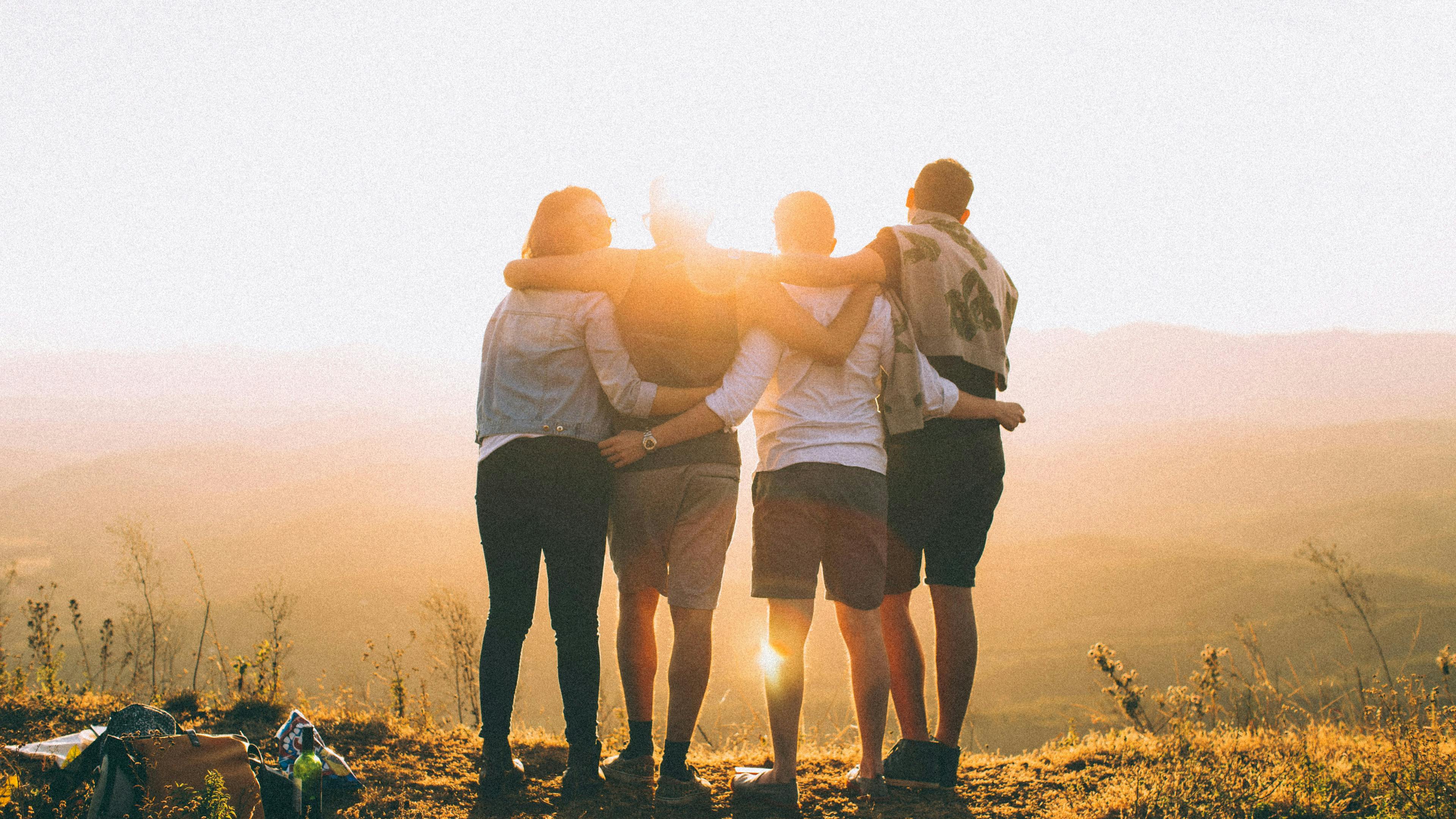 Four friends stand with their arms around each other's shoulders, looking out over a vast mountain valley at sunset. The bright sun creates a warm glow and silhouettes the group against the hazy horizon.