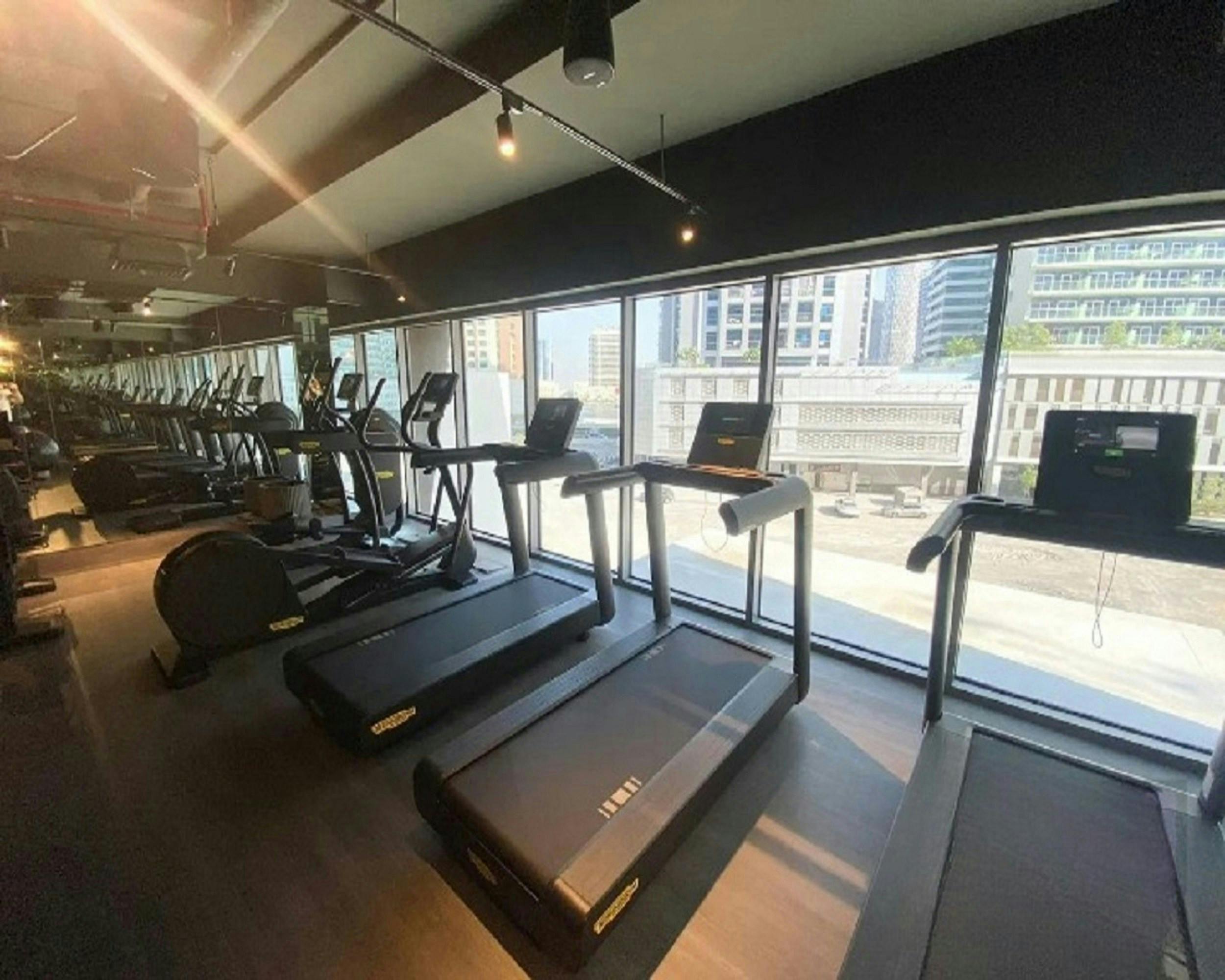 A bright, modern commercial gym space featuring a row of high-end Technogym treadmills facing large floor-to-ceiling windows with a city view.