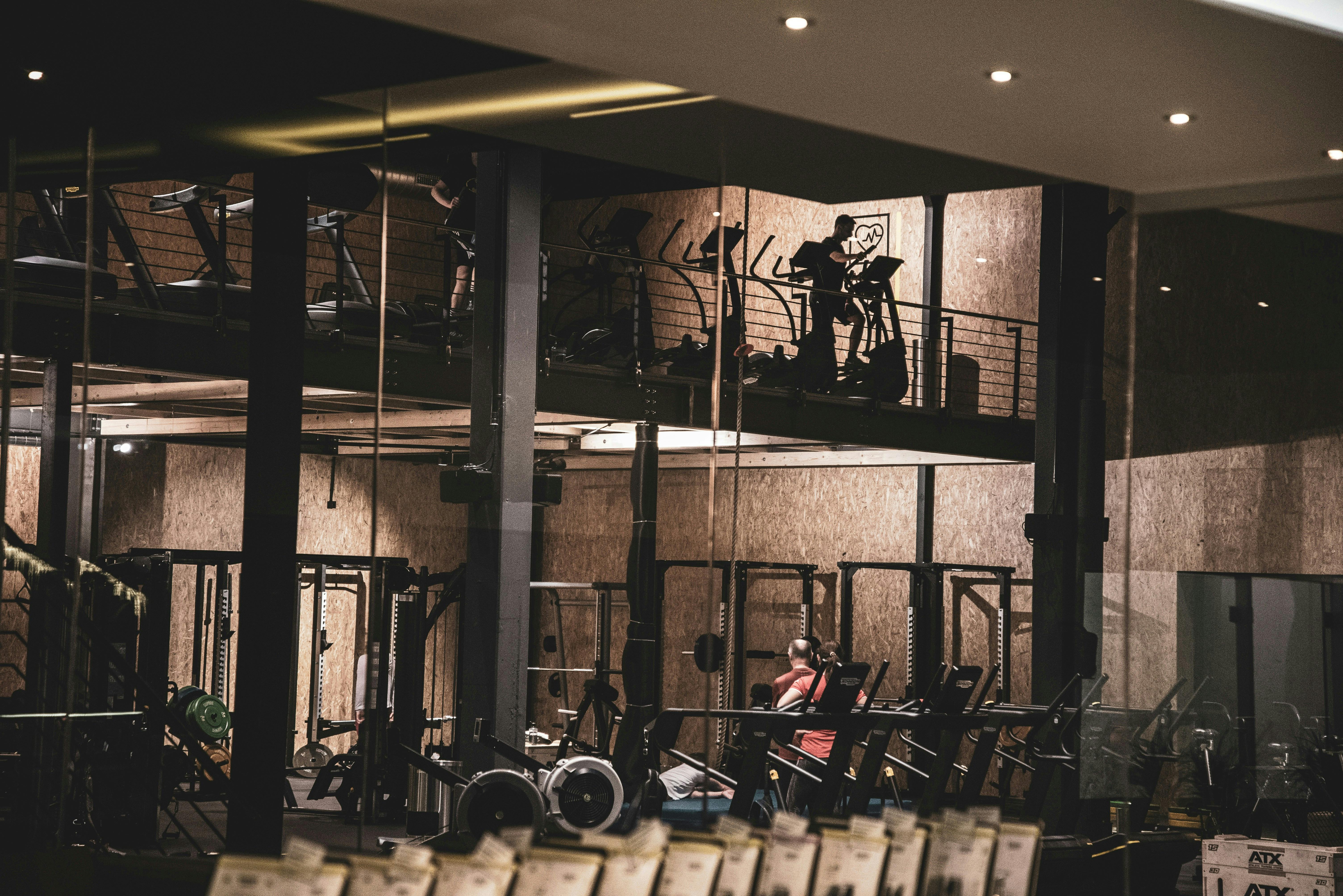 A moody, industrial-style boutique gym featuring a two-story layout with heavy-duty squat racks on the ground floor and cardio equipment on a mezzanine level.