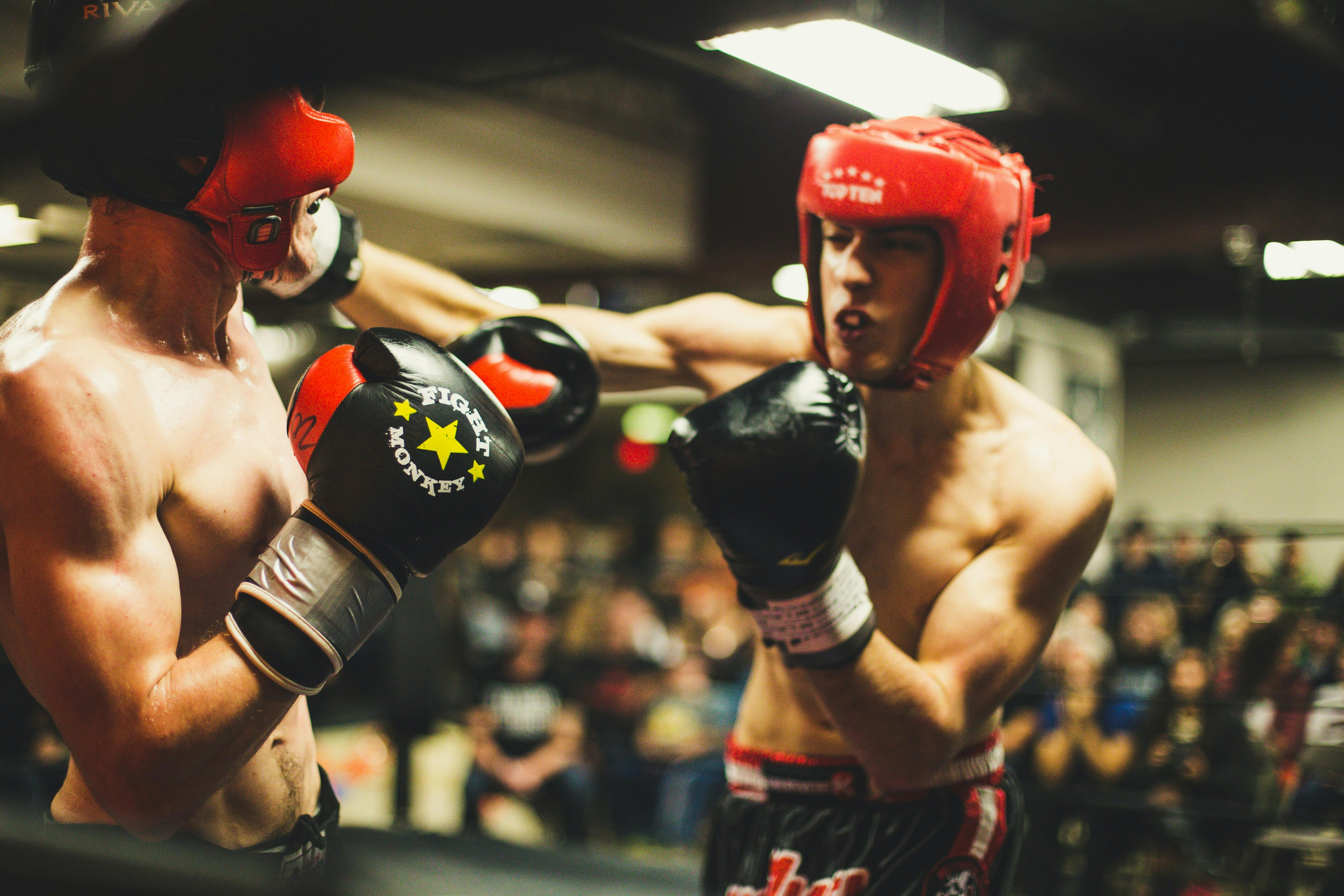 An intense close-up of two amateur boxers mid-sparring session, wearing protective headgear and gloves, during a competitive match with a crowd in the background.