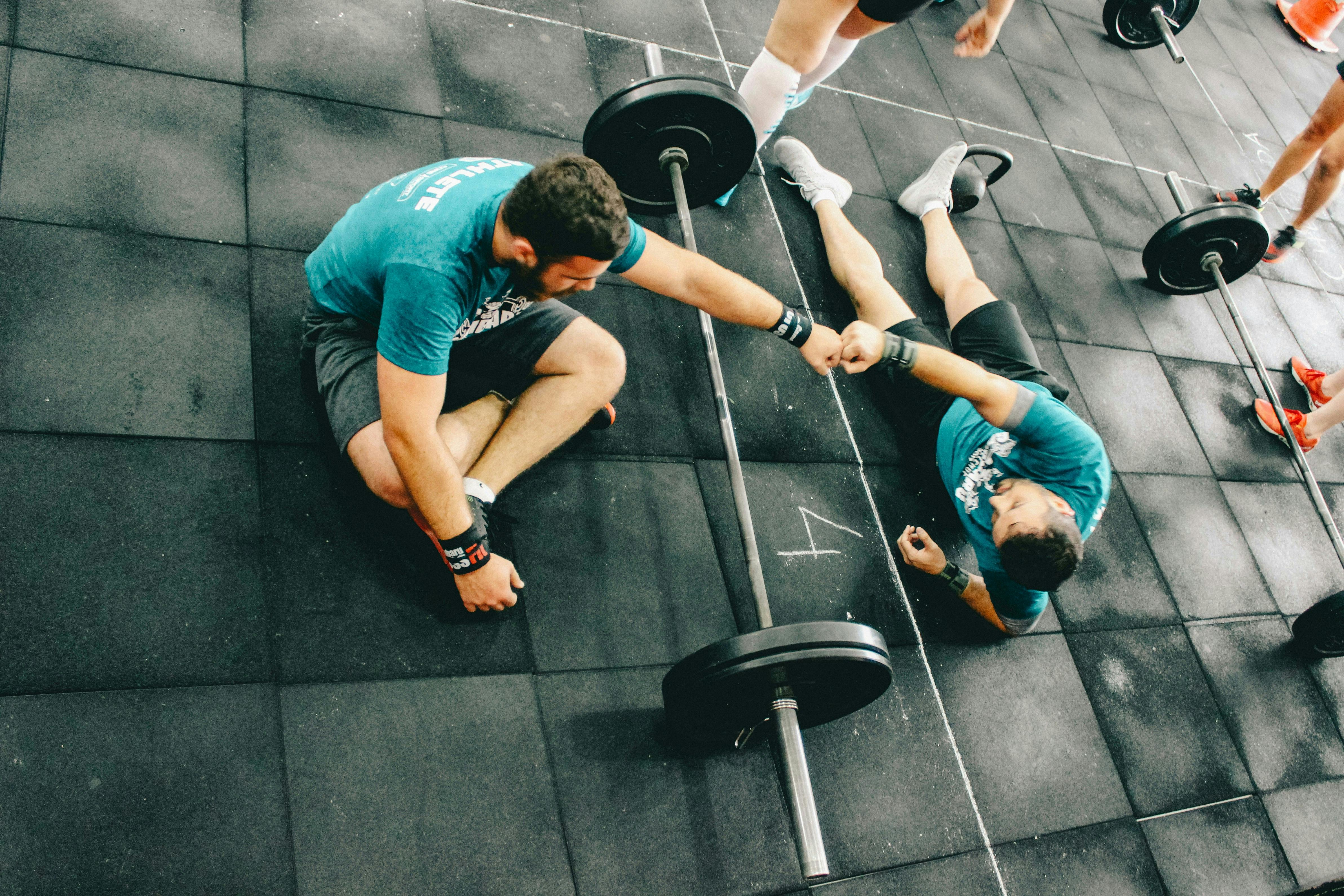 Two CrossFit athletes sharing a "fist bump" of mutual respect while resting on a black rubber-matted floor next to loaded barbells after a grueling workout.