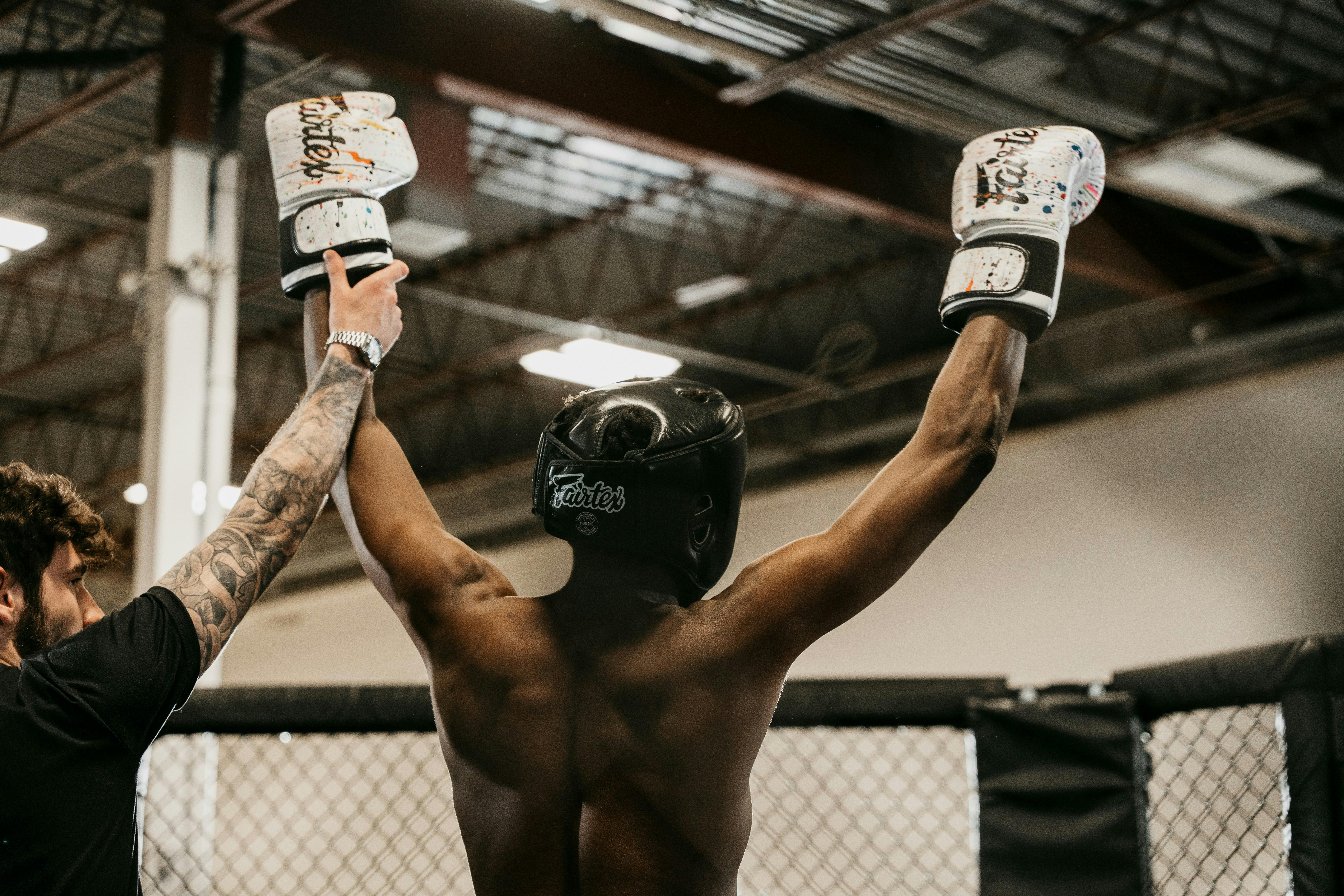 A triumphant mixed martial arts fighter with their back to the camera, raising their arms in victory inside an octagon cage while the referee holds their wrist.