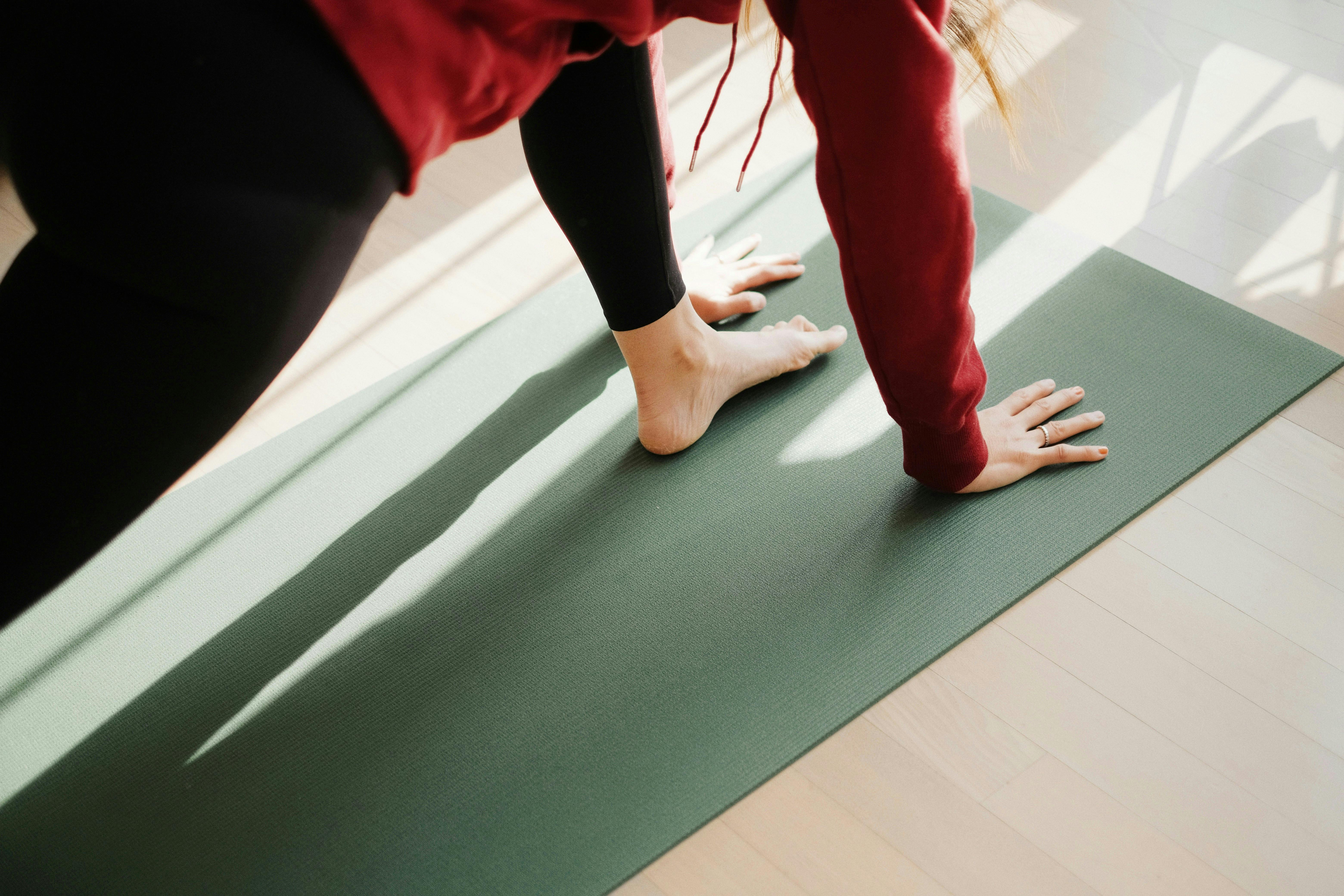A minimalist, top-down view of a person in a yoga pose on a sage green mat, with soft natural sunlight streaming across the light wood floor.