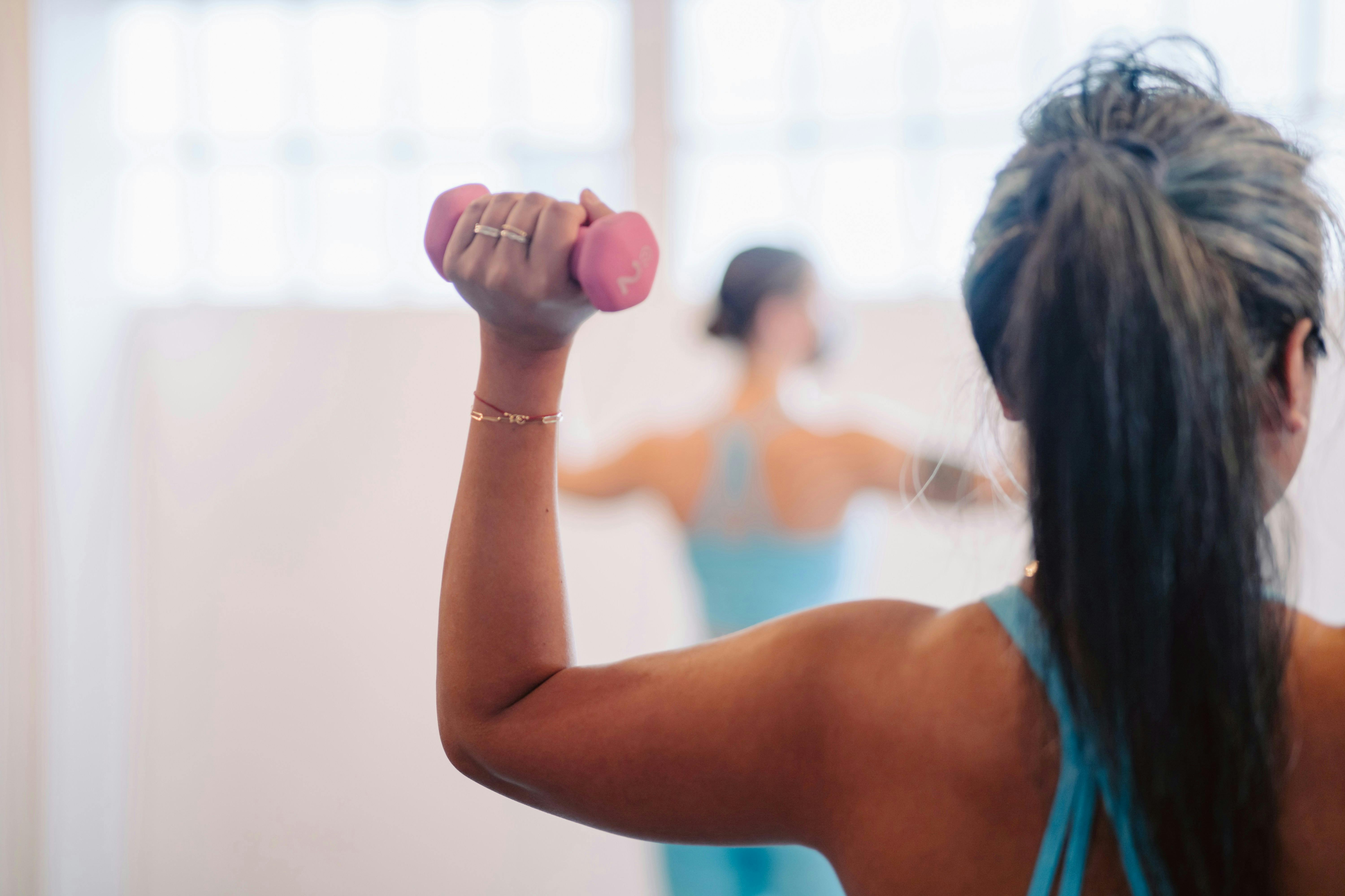 A focused shot from behind a woman in a barre class, lifting a small pink dumbbell to tone her arms in a bright, airy studio setting.