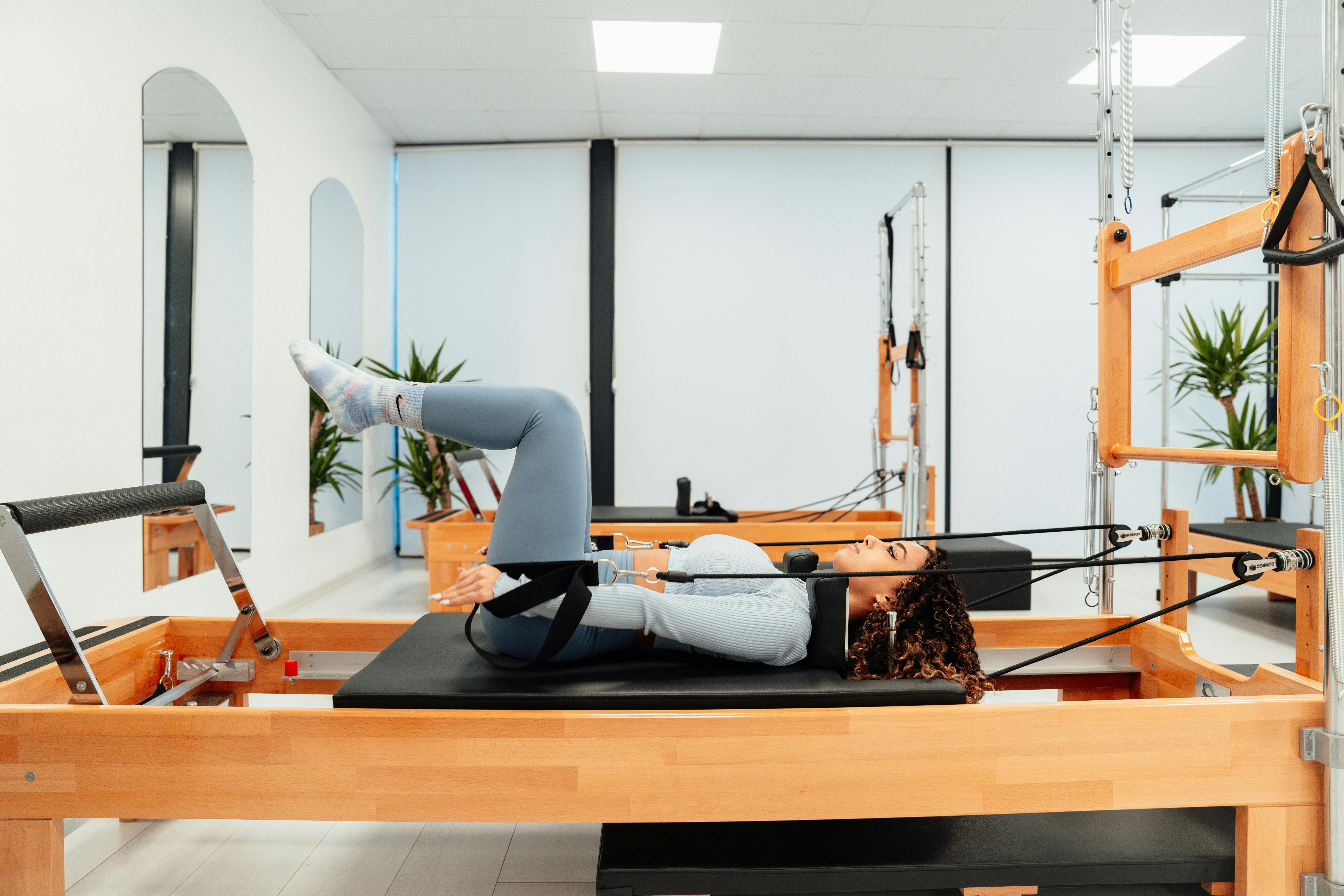 A woman performing a controlled core exercise on a wooden Pilates reformer machine in a clean, professional studio with large mirrors.