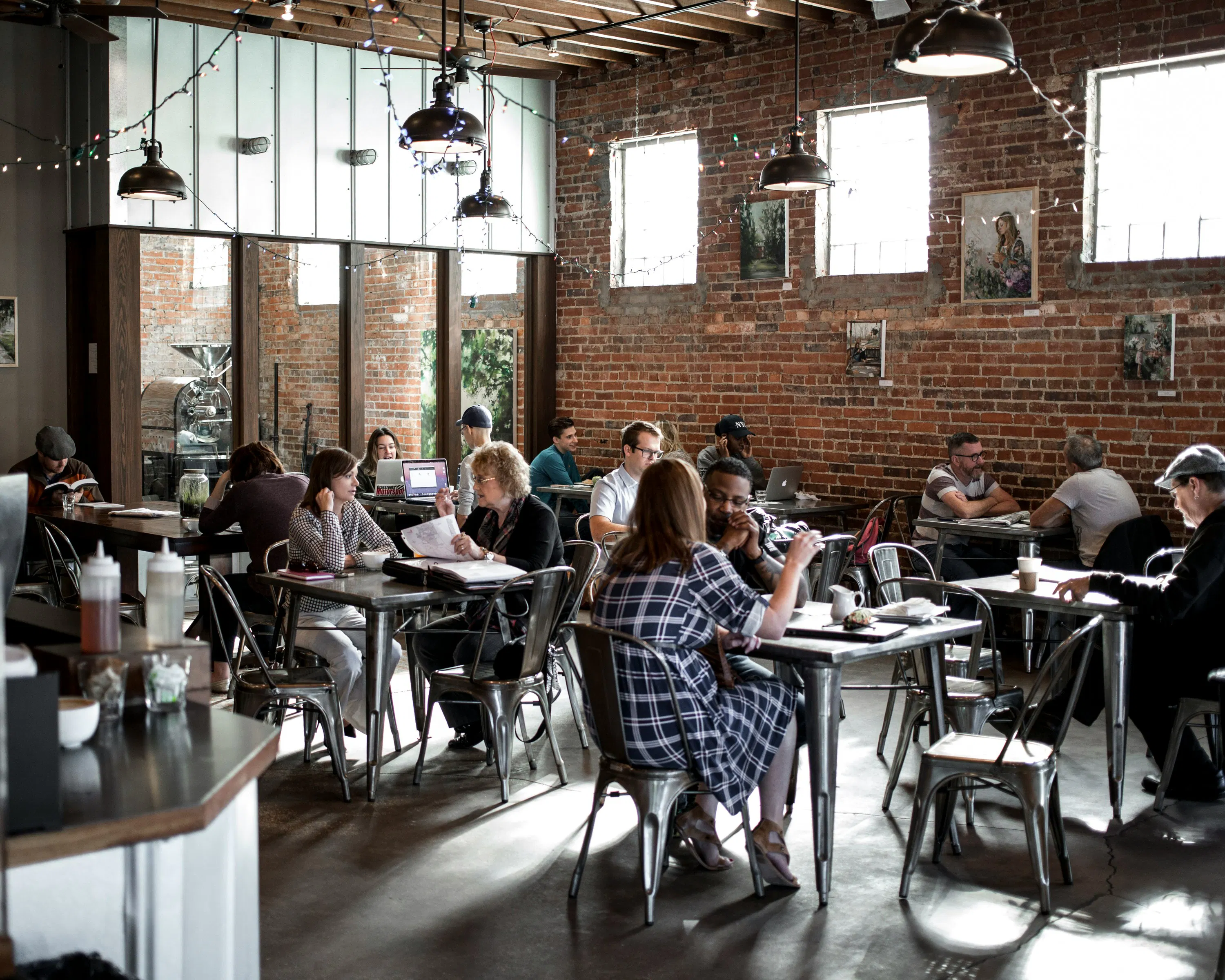 A bustling, industrial-style coffee shop filled with people working on laptops and chatting at metal tables against a rustic brick wall.