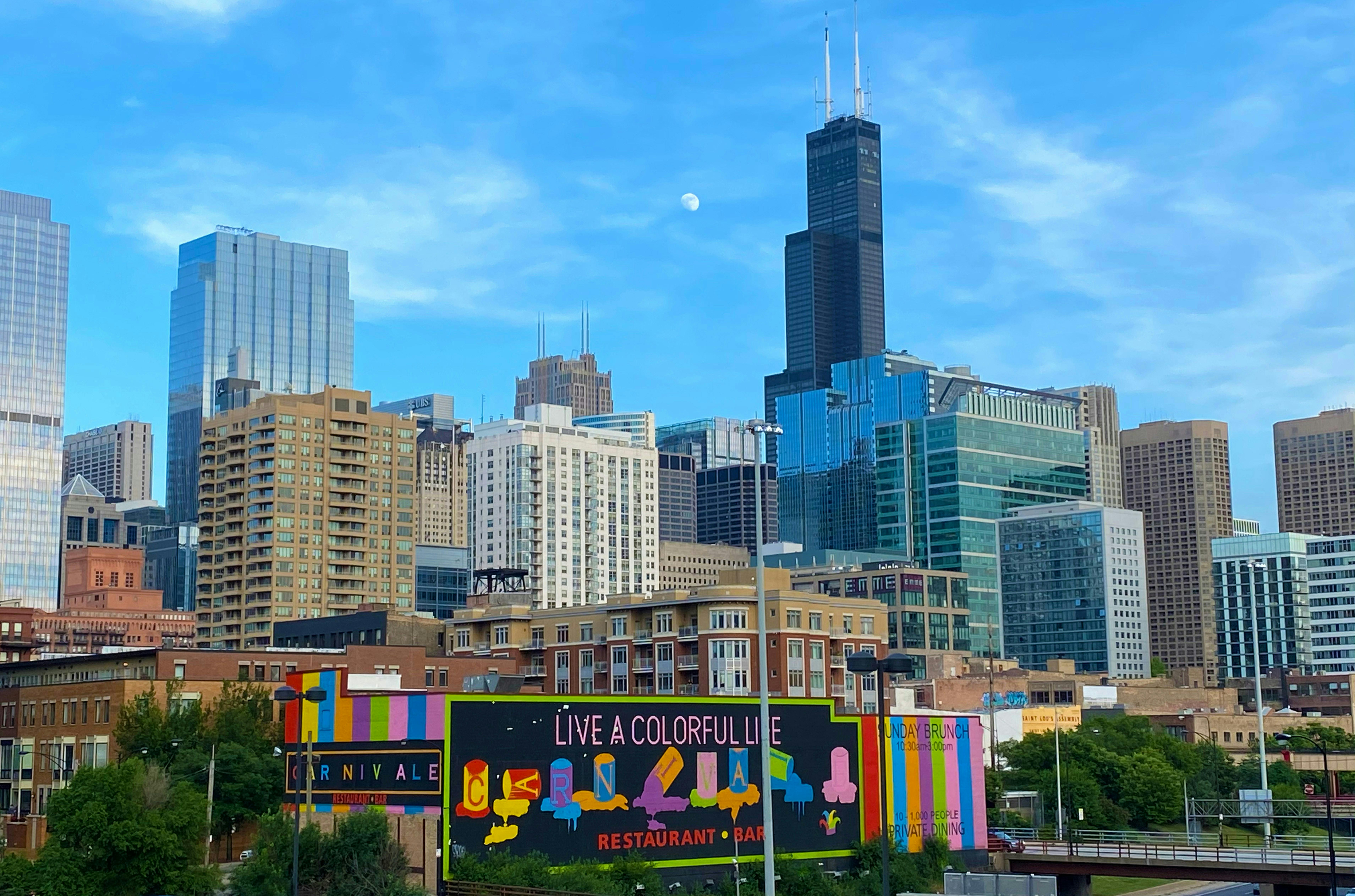 A cityscape of Chicago’s West Loop featuring the colorful "Live a Colorful Life" mural on the side of the Carnivale restaurant. In the background, the Willis Tower and various skyscrapers stand tall under a clear blue sky with a visible daytime moon.