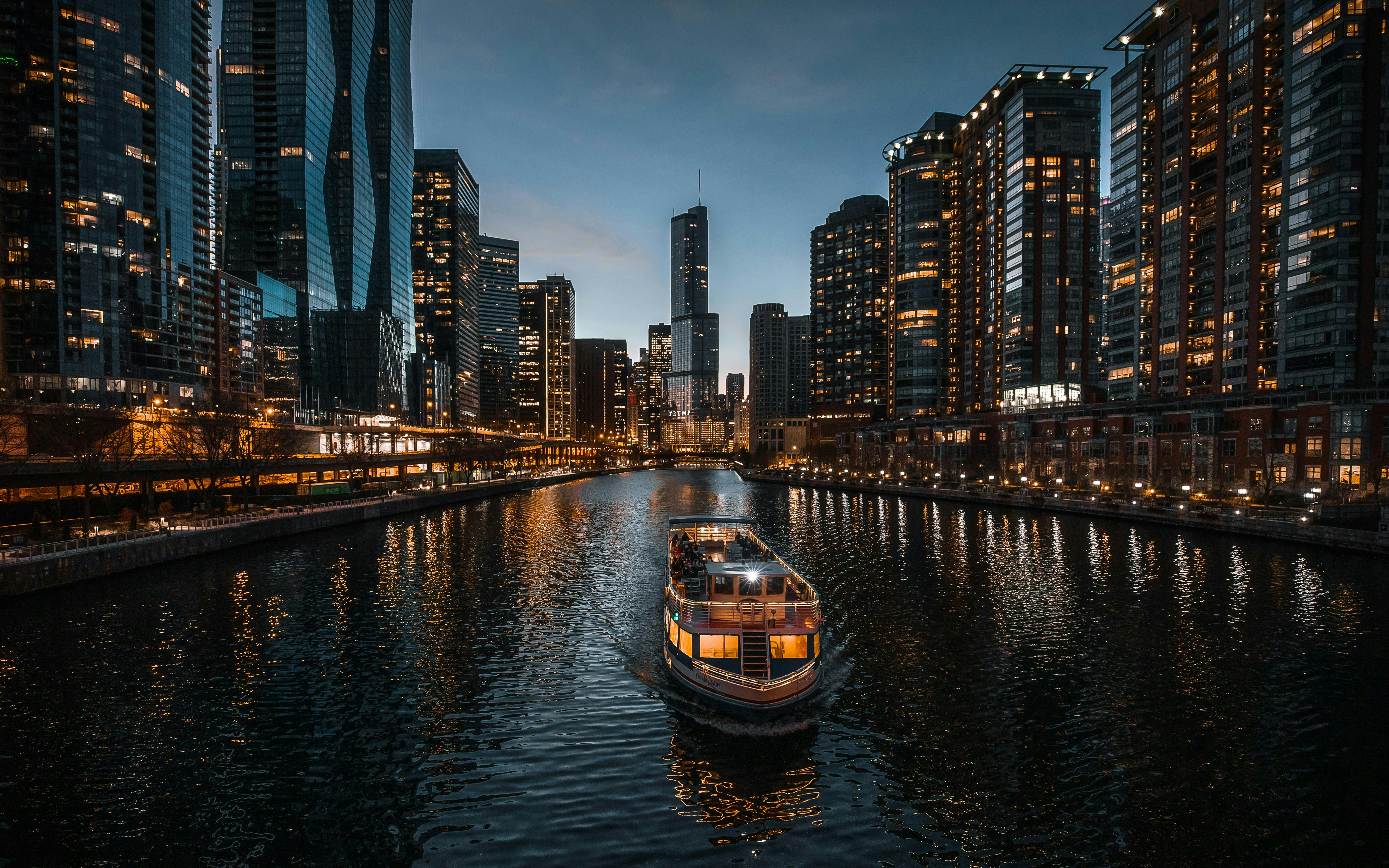 A night view of a tour boat illuminated with warm lights traveling down the Chicago River. The water reflects the glowing lights of the surrounding modern skyscrapers and high-rise apartments under a deep blue twilight sky.