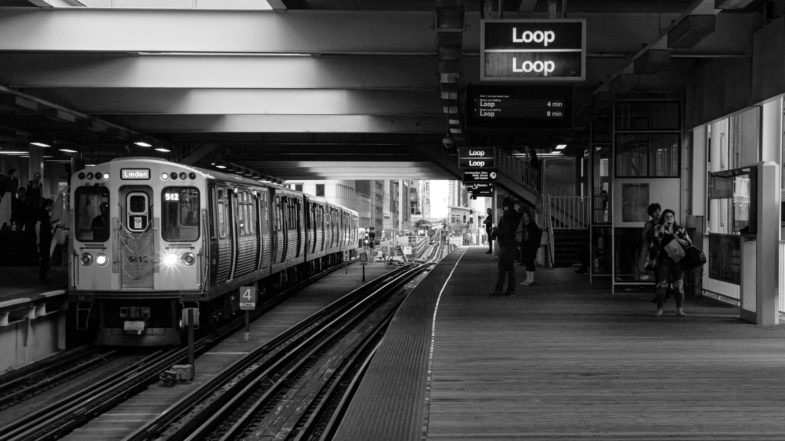 A black and white photograph of an elevated CTA train platform in Chicago. A train labeled "Linden" is stopped at the platform on the left, while commuters wait near digital signs displaying "Loop." The perspective looks down the tracks toward the open city street in the distance.