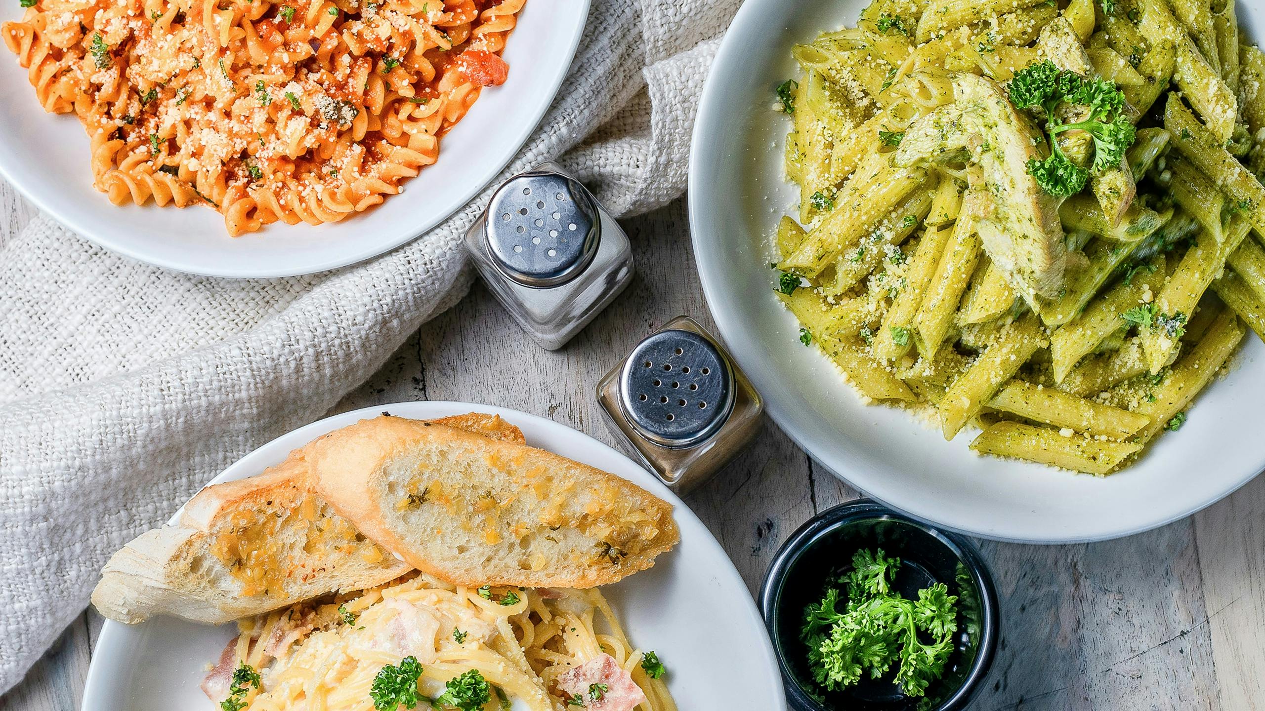 A top-down view of three different Italian pasta dishes and sliced bread served on a rustic gray wooden table