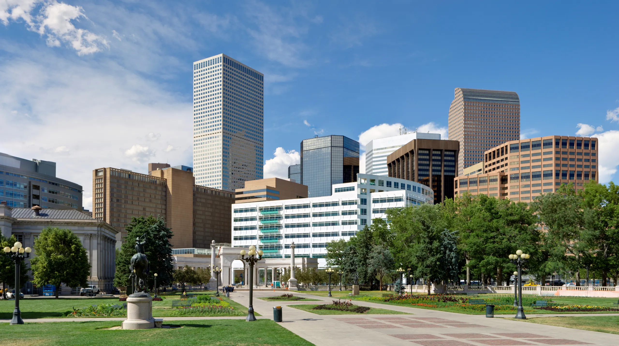 A wide view of a city park in the Golden Triangle with green lawns, a monument, and a background of modern skyscrapers.