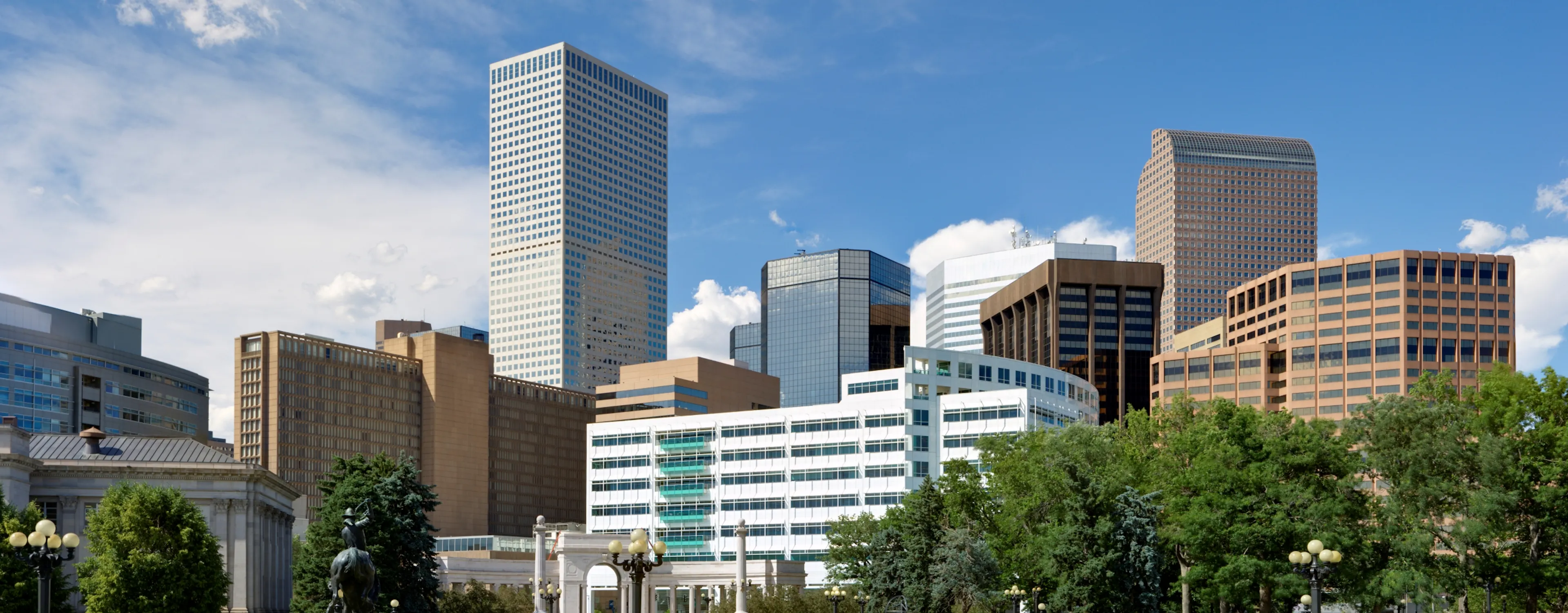 A wide view of a city park in the Golden Triangle with green lawns, a monument, and a background of modern skyscrapers.