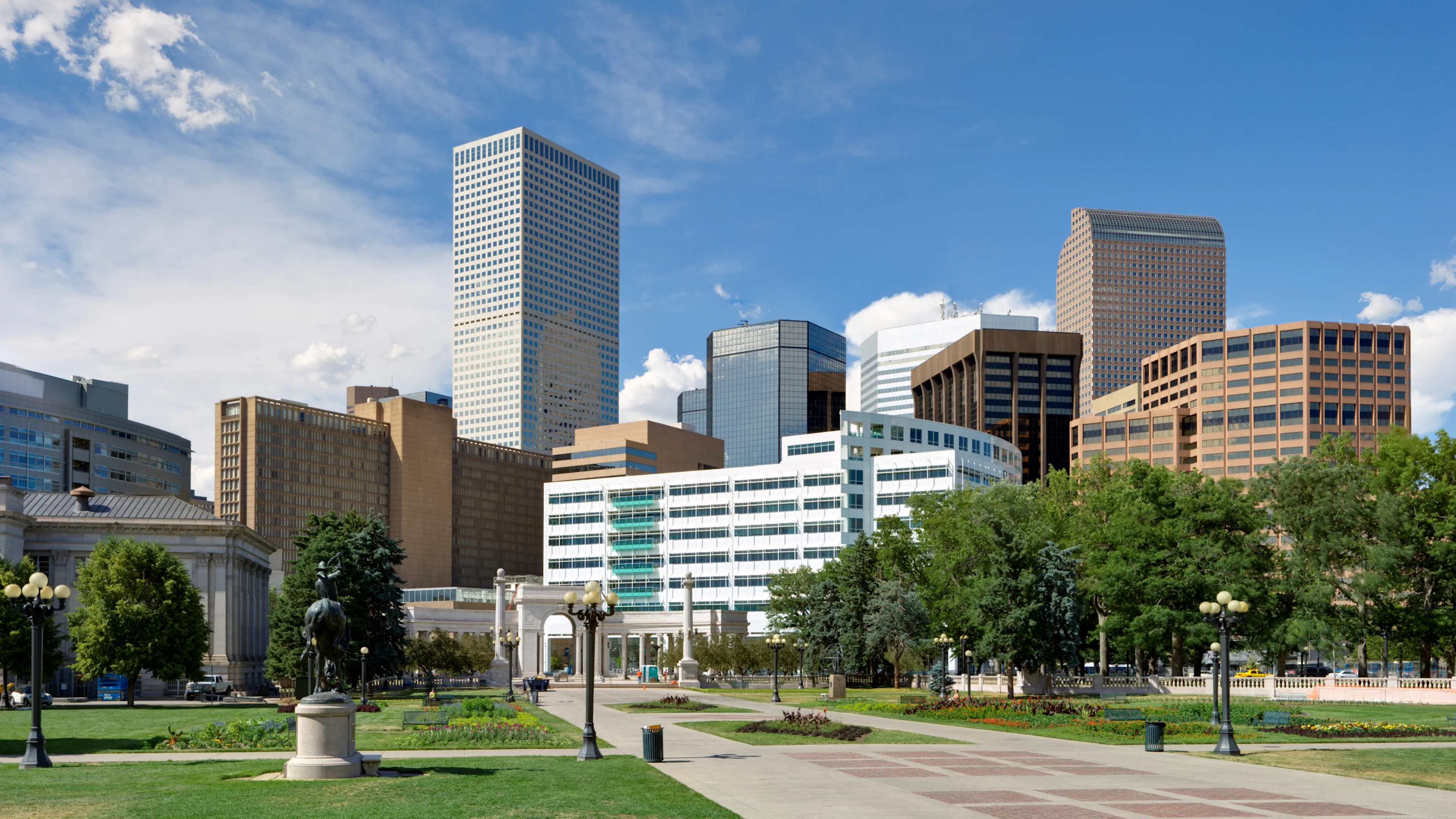 A wide view of a city park in the Golden Triangle with green lawns, a monument, and a background of modern skyscrapers.