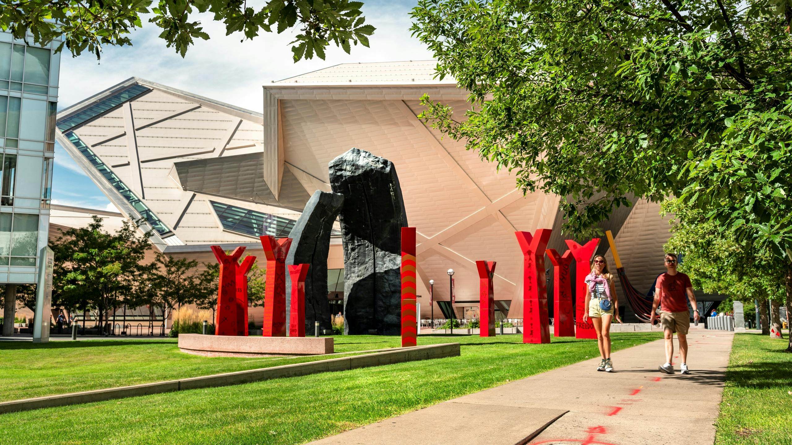 People walking past modern red sculptures and unique architectural buildings in a sunny plaza in the Golden Triangle.