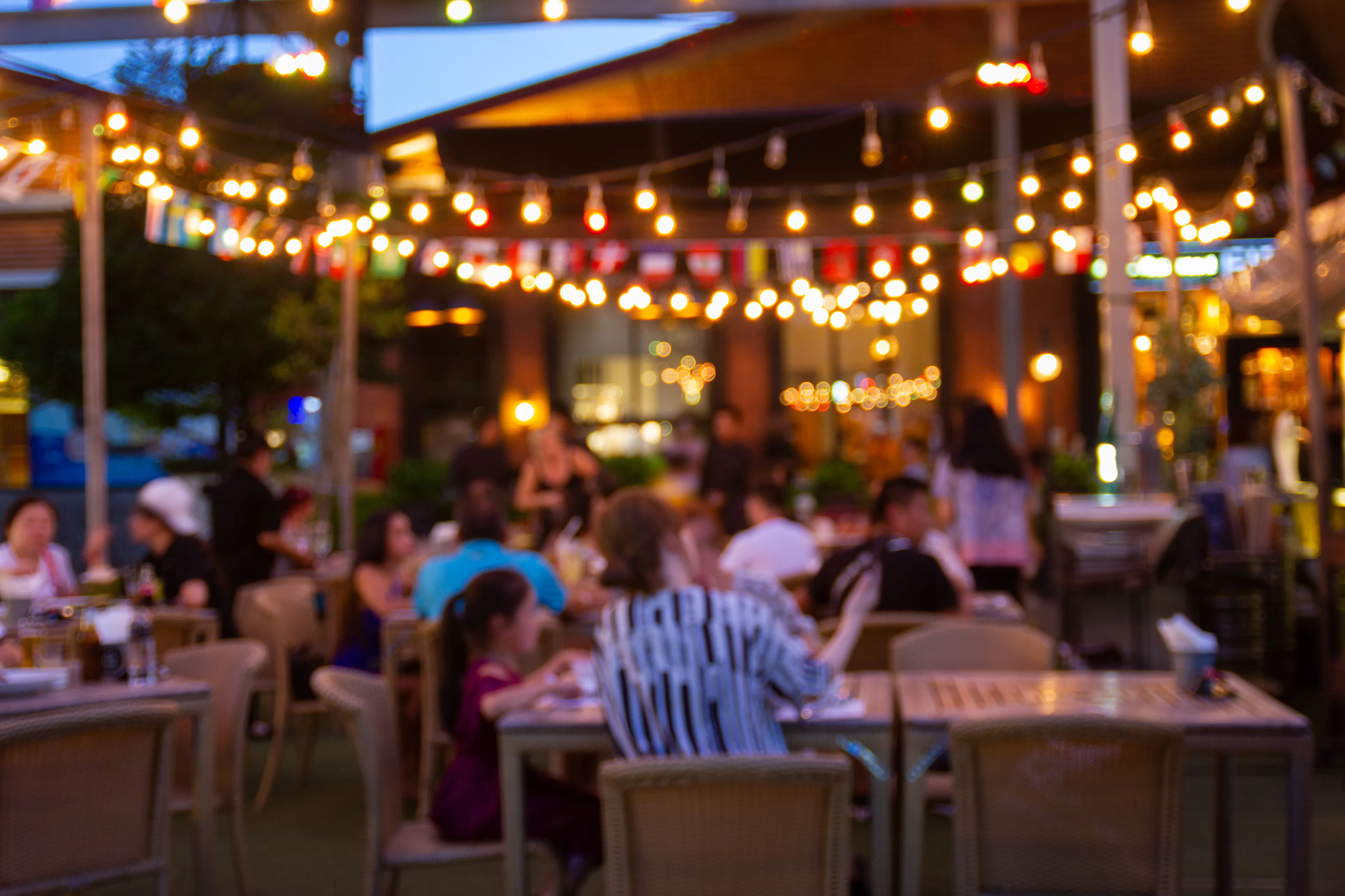 Blurred evening view of a lively outdoor restaurant patio featuring glowing string lights, international flags, and local dining