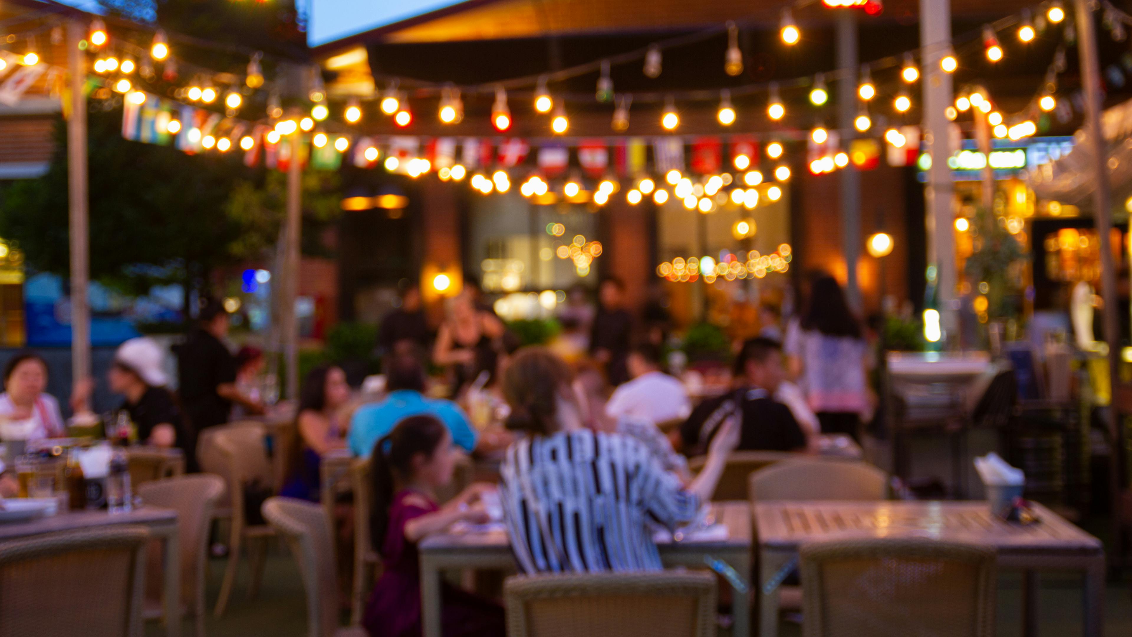 Blurred evening view of a lively outdoor restaurant patio featuring glowing string lights, international flags, and local dining