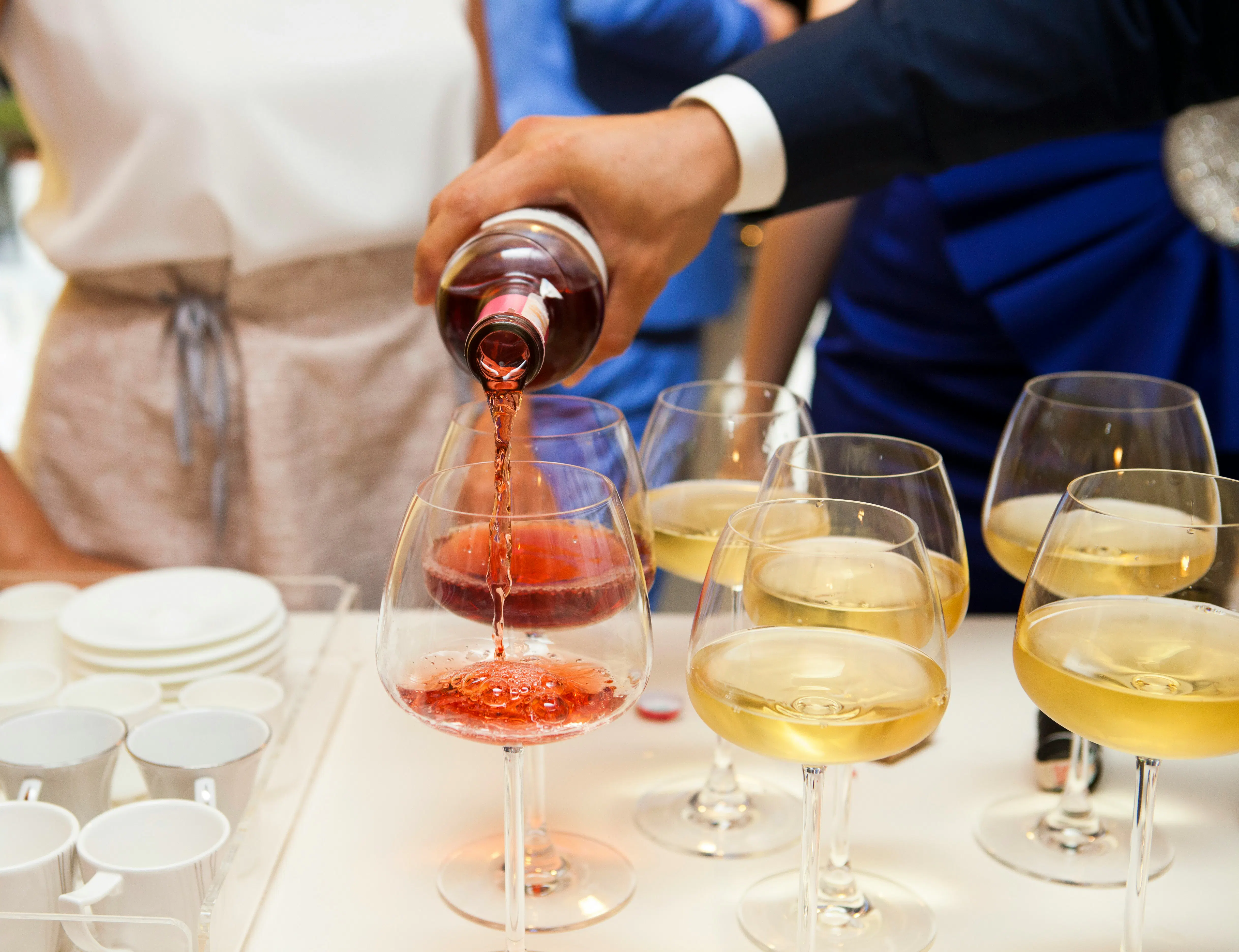 A person in a dark suit pours a vibrant rosé wine into a glass during a tasting event. Several other glasses filled with white wine are arranged on the white tabletop, next to a tray of small white ceramic cups.

