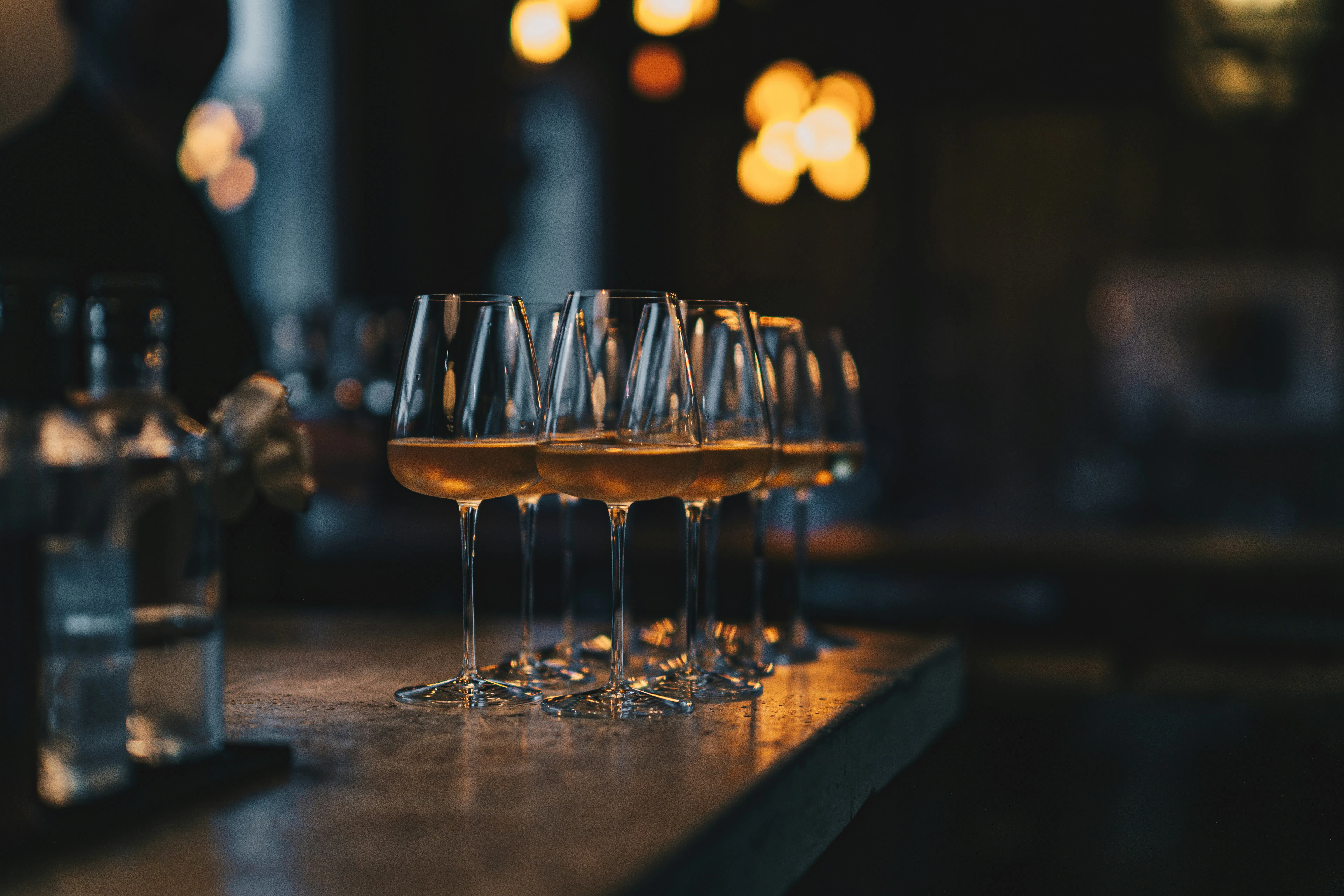 A row of wine glasses filled with white wine stands on a textured concrete bar. The lighting is moody and dark, with warm, glowing bokeh lights in the background creating a sophisticated atmosphere.

