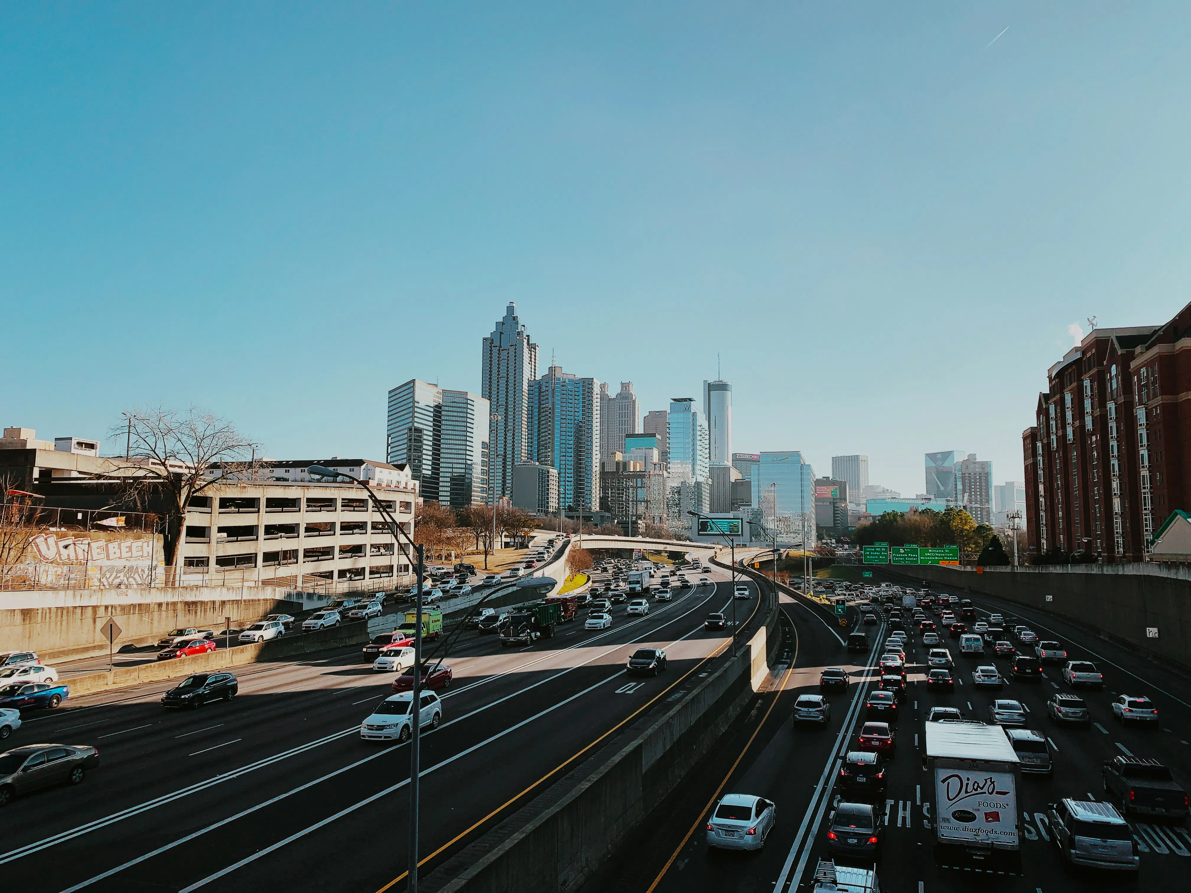 A wide-angle shot of a multi-lane highway leading toward the Atlanta city skyline under a clear blue sky. Heavy traffic fills the lanes in both directions. The urban backdrop features prominent skyscrapers, including the Westin Peachtree Plaza and the Georgia-Pacific Center.