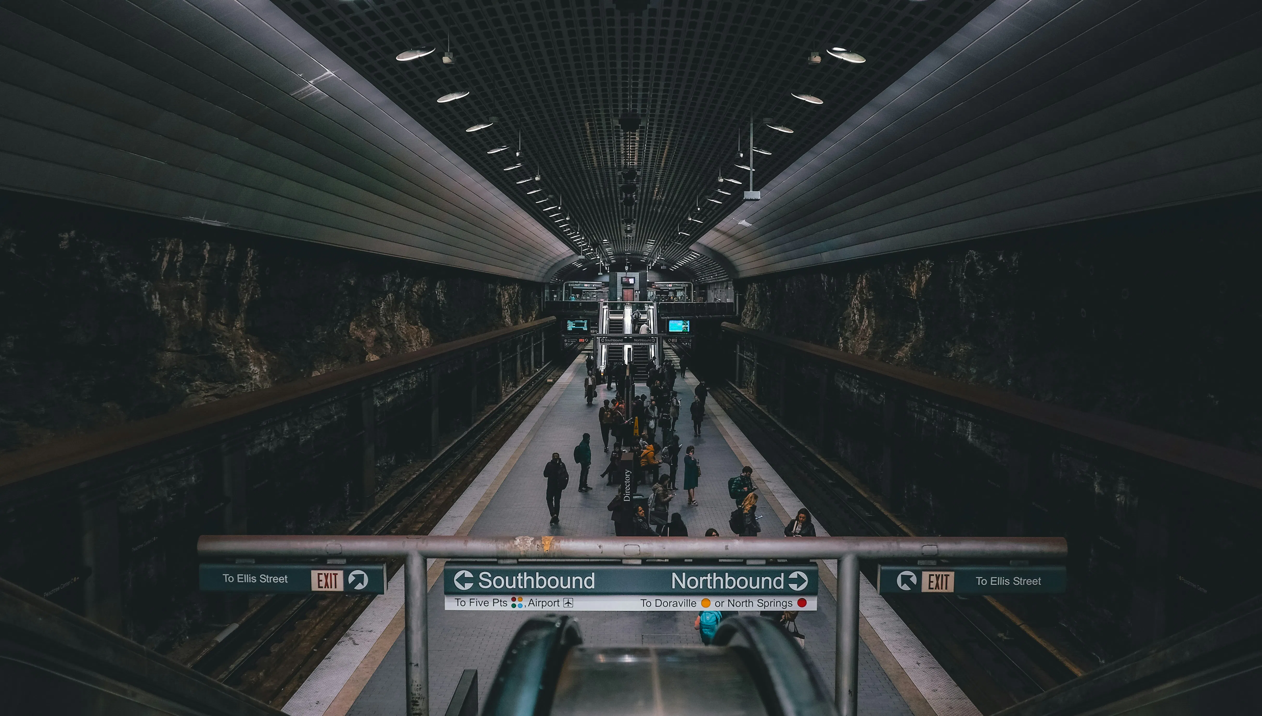 A symmetrical, low-angle shot of a MARTA subway station platform from the top of an escalator. Commuters stand along the platform under a vaulted, industrial ceiling with recessed lighting. Signs hanging from a metal rail indicate "Southbound to Airport" and "Northbound to Doraville or North Springs."