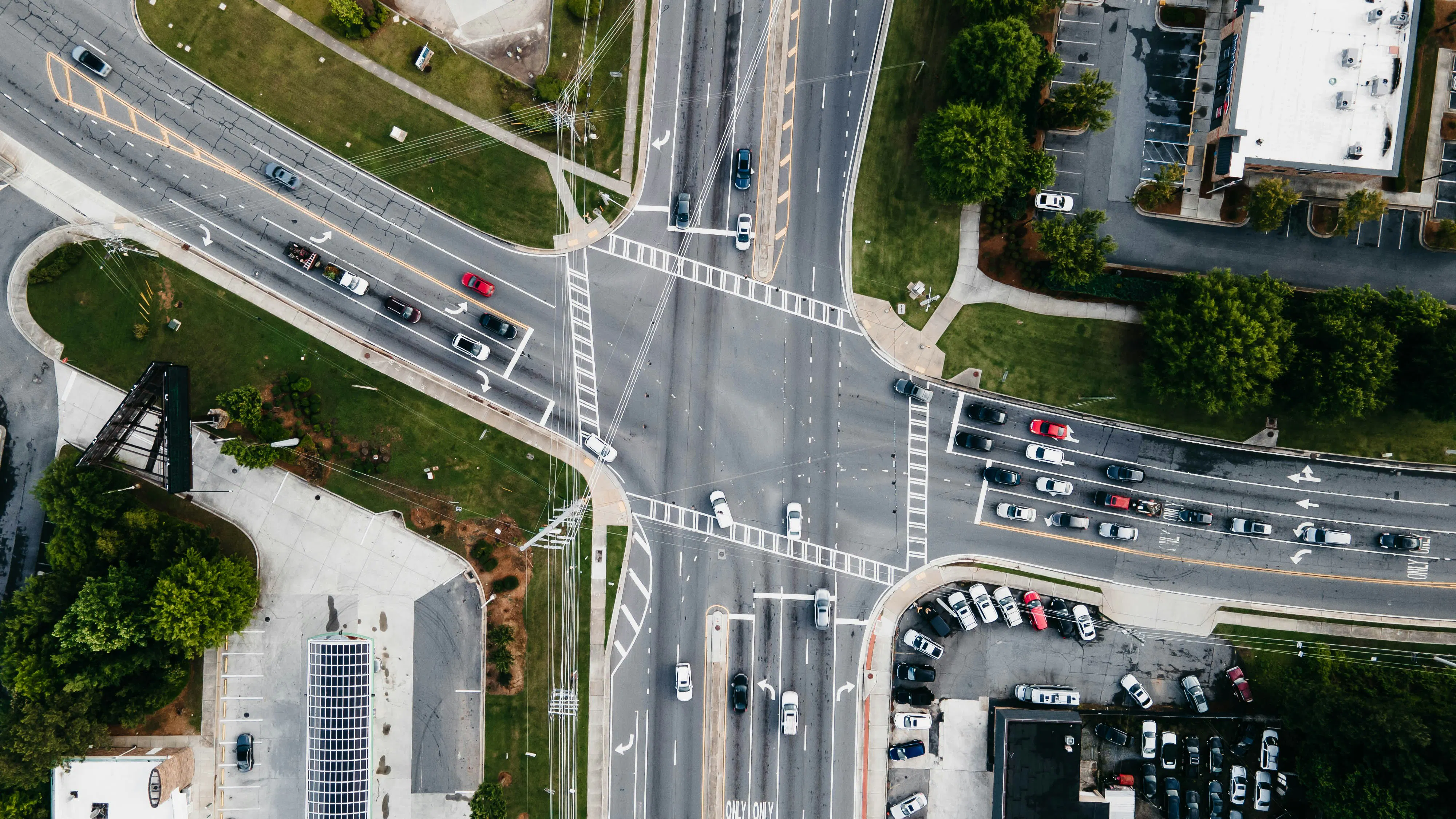 An overhead, bird's-eye view of a large, complex four-way road intersection. Multiple lanes are filled with cars, and clear white crosswalks and directional arrows are painted on the asphalt. The surrounding areas include green grass, parking lots, and buildings with solar panels.