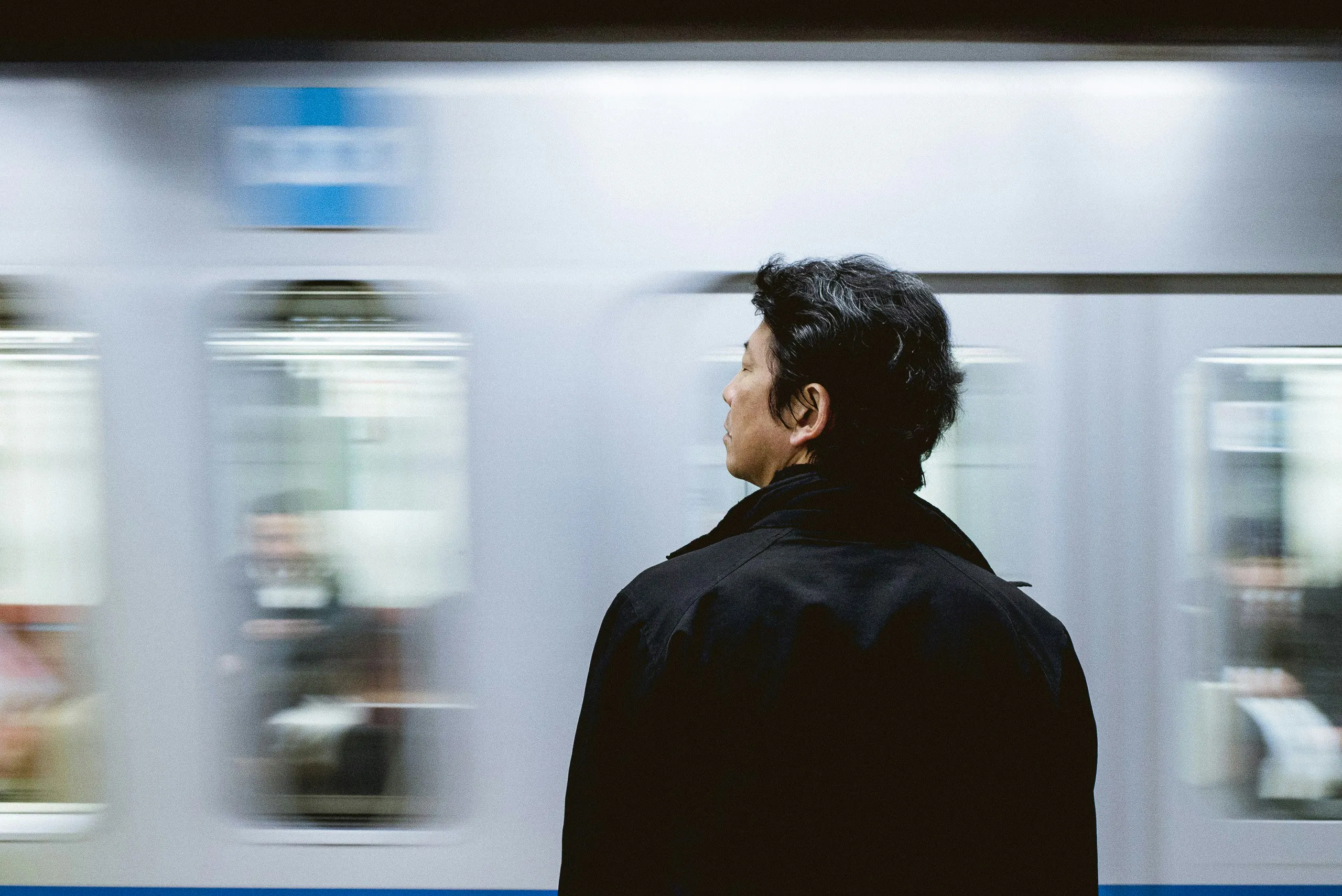 A candid, close-up profile of a man with dark hair wearing a black jacket, standing on a subway platform. In the background, a silver train is blurred in motion, creating a sense of speed and urban energy.