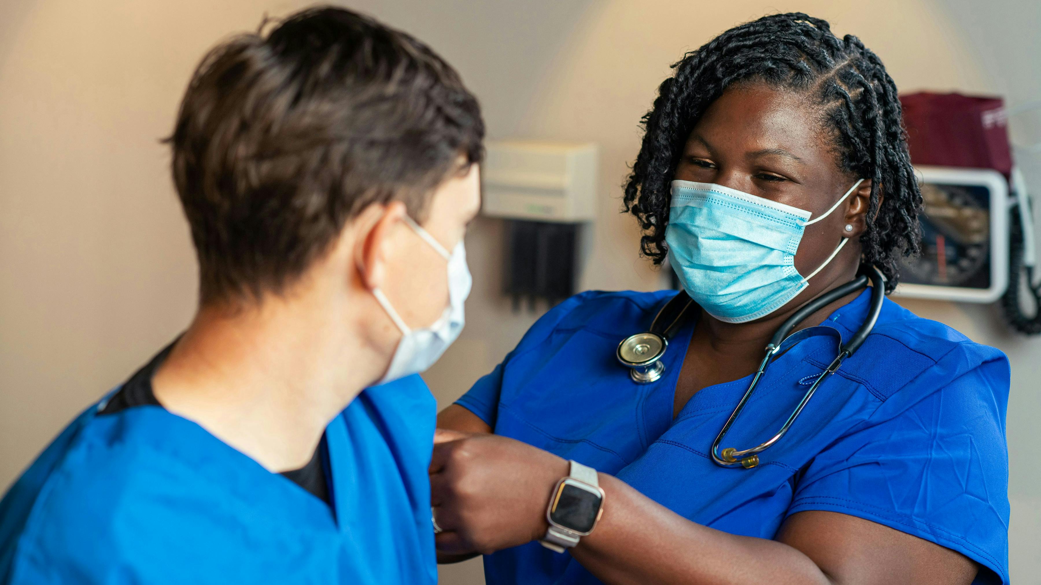 A close-up of a healthcare professional with braided hair wearing blue scrubs and a surgical mask. She is focused on a patient (whose back is to the camera) while performing a medical procedure, likely administering a vaccination or checking a bandage. A stethoscope hangs around her neck.