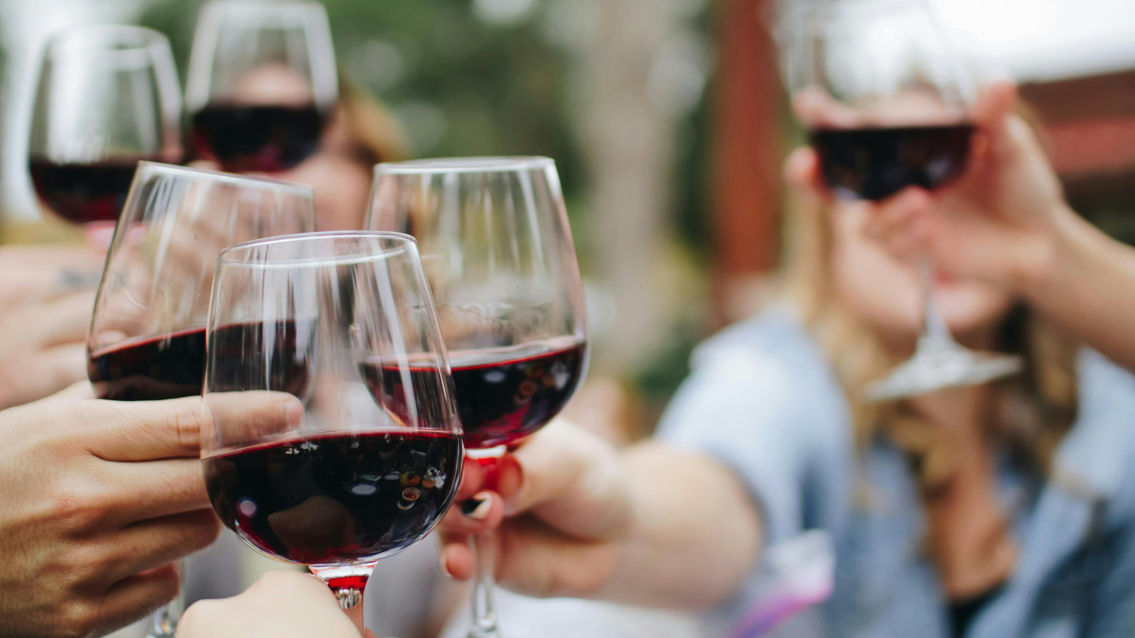 A joyful, outdoor scene of multiple people raising six glasses of red wine in a celebratory toast. The focus is sharp on the wine glasses in the foreground, with the smiling faces of the friends blurred in the background.