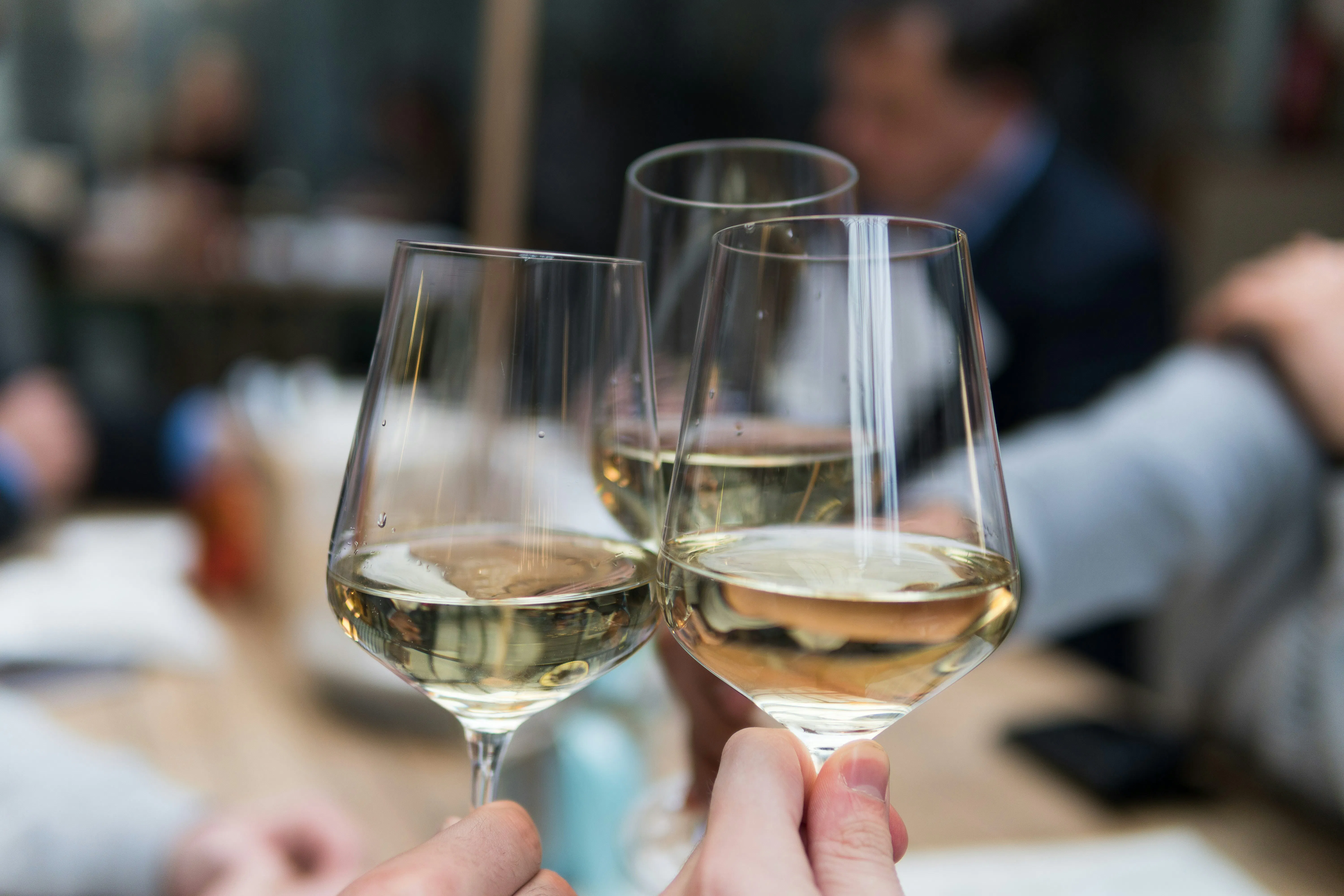 A close-up of three glasses of chilled white wine being clinked together in a toast. The background is softly blurred, showing an outdoor or well-lit restaurant patio setting.