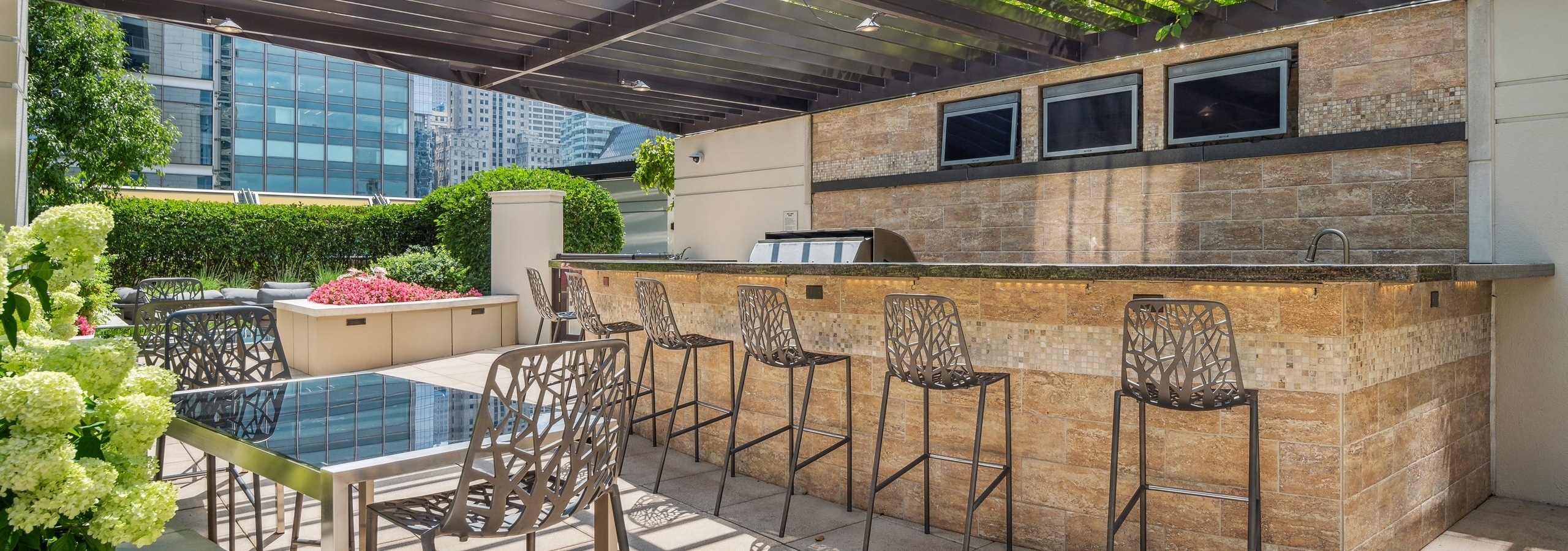 AMLI River North outdoor kitchen with bar top and 6 barstools with TVs and grill with dining tables shaded by metal canopy