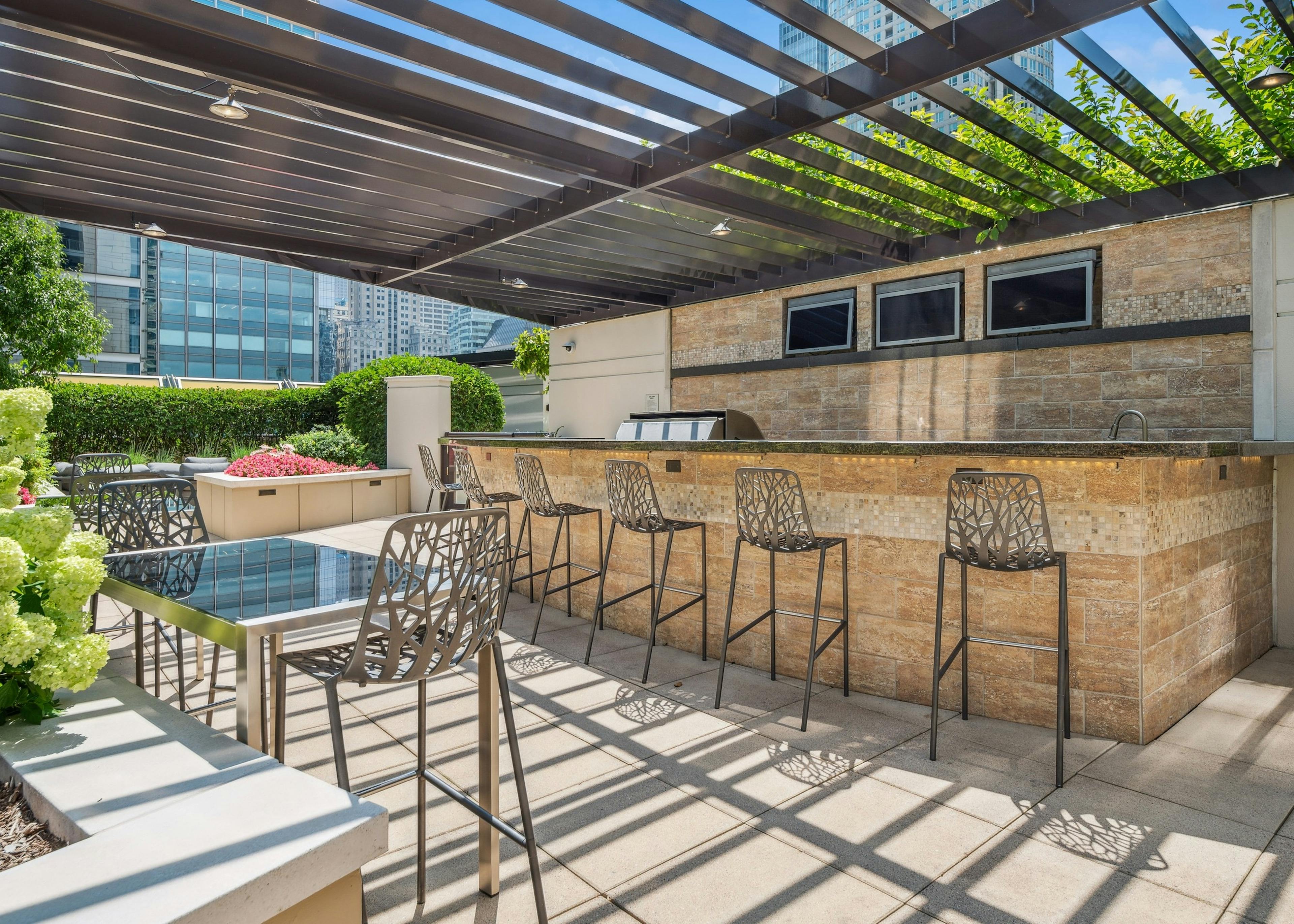 AMLI River North outdoor kitchen with bar top and 6 barstools with TVs and grill with dining tables shaded by metal canopy