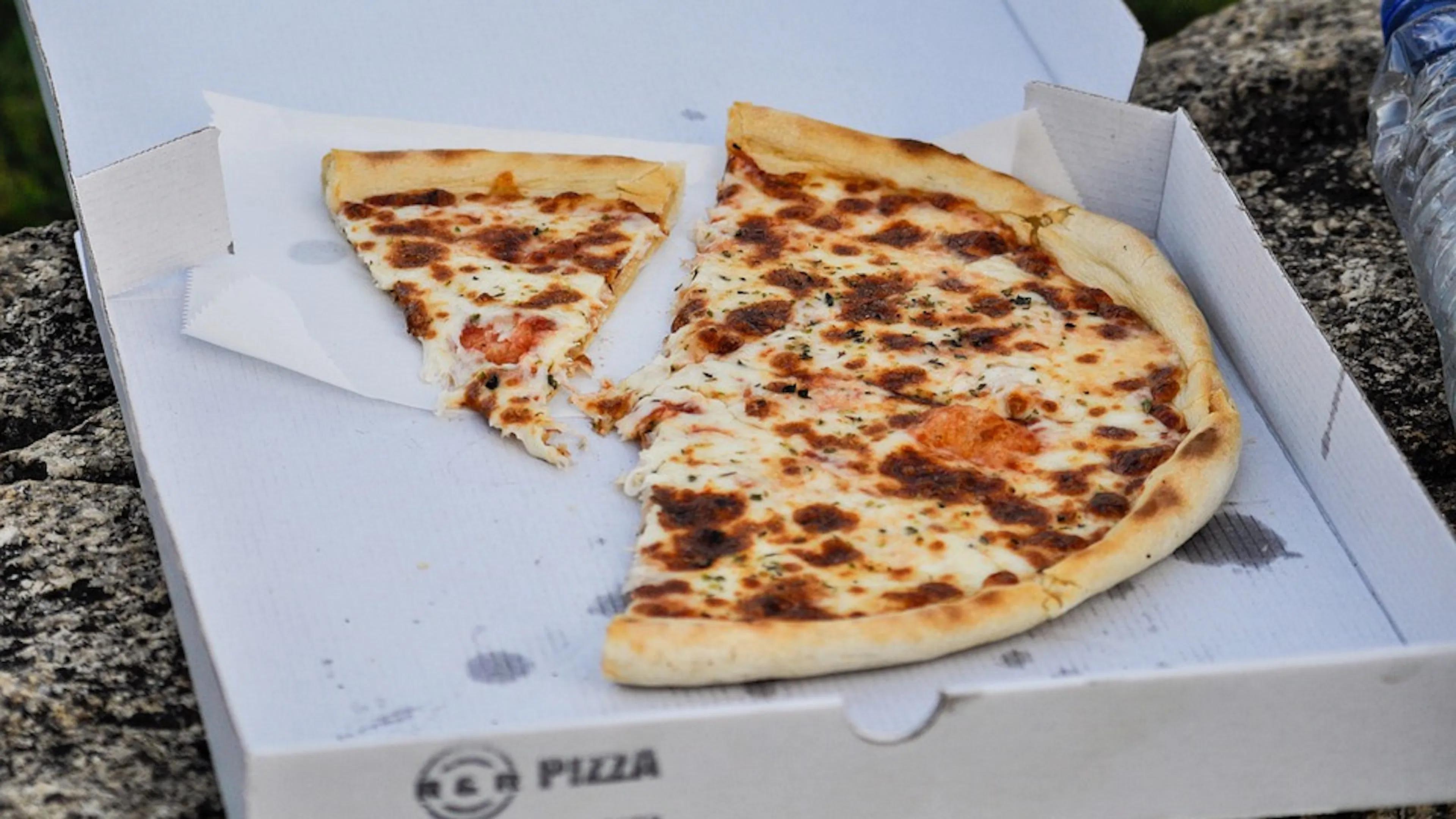 Half a plain cheese pizza in a white cardboard pizza box set on a gray rock
