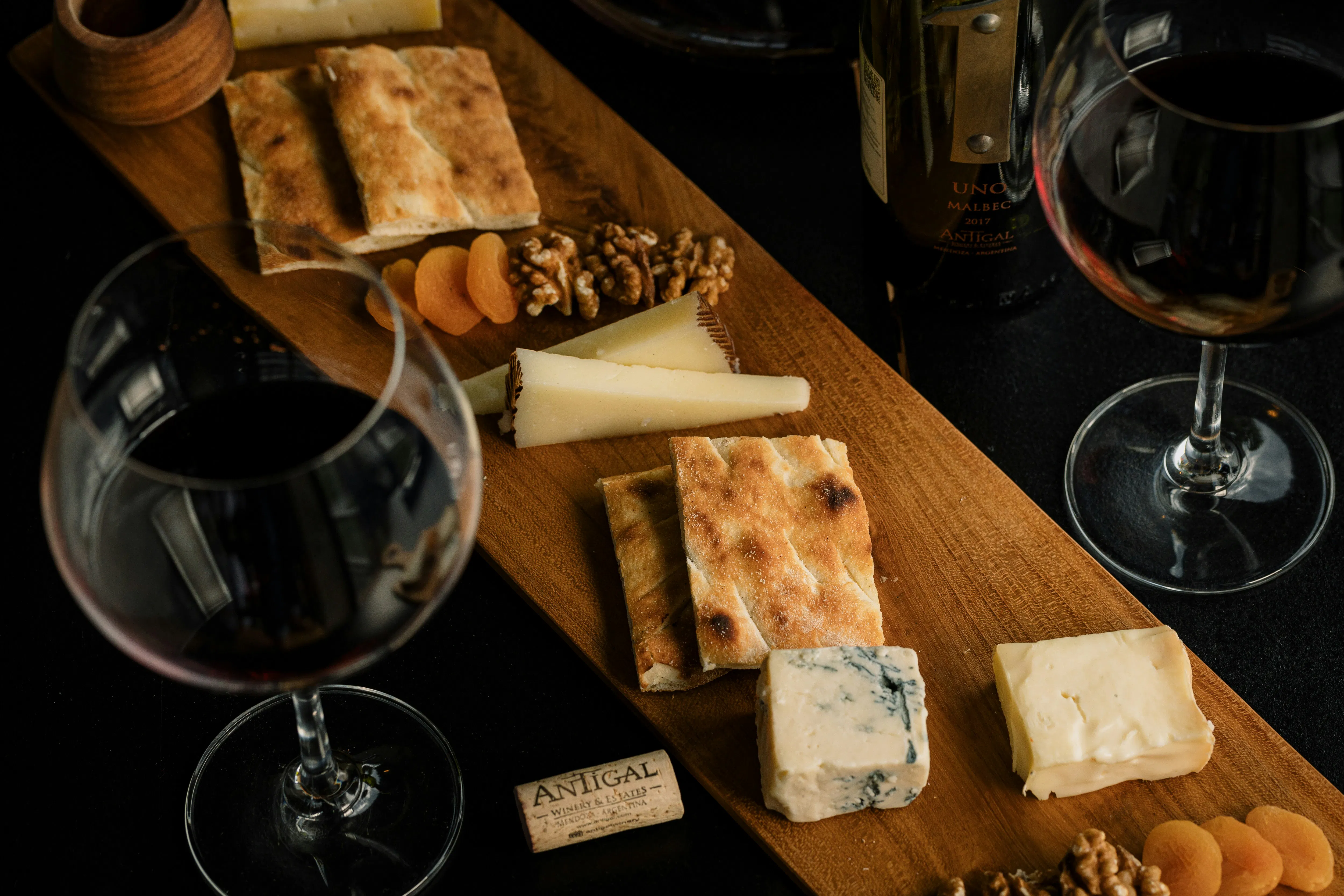 A top-down, close-up shot of a long wooden charcuterie board featuring various cheeses, including wedges of Manchego, a block of blue cheese, and a creamy soft cheese. It is garnished with dried apricots, walnuts, and toasted flatbread, accompanied by two glasses of red wine and a bottle of Malbec.