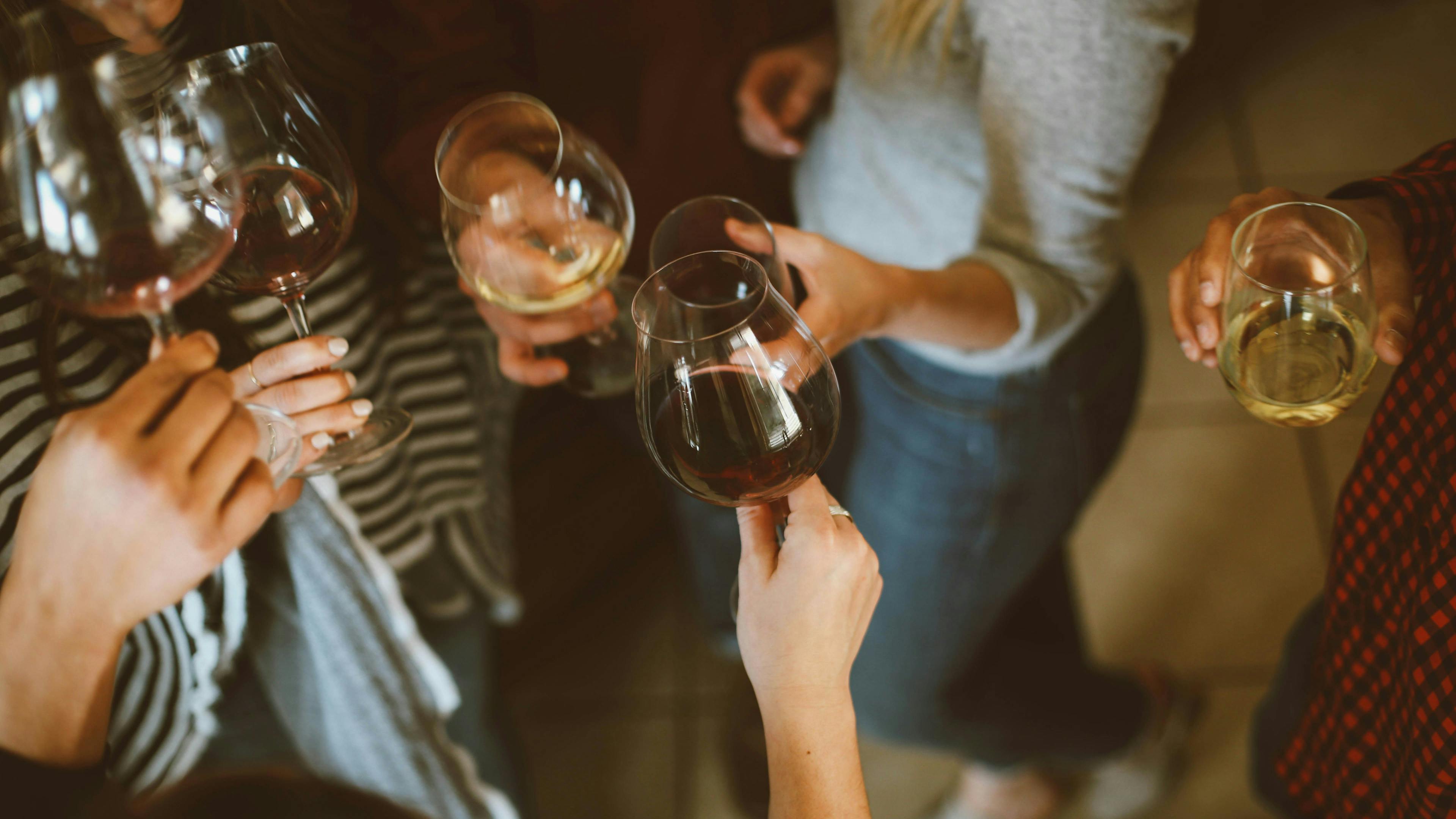 An overhead, lifestyle shot of a group of friends standing together and toasting with a mix of red and white wine. The warm, casual atmosphere shows the group in comfortable clothing like sweaters and jeans.