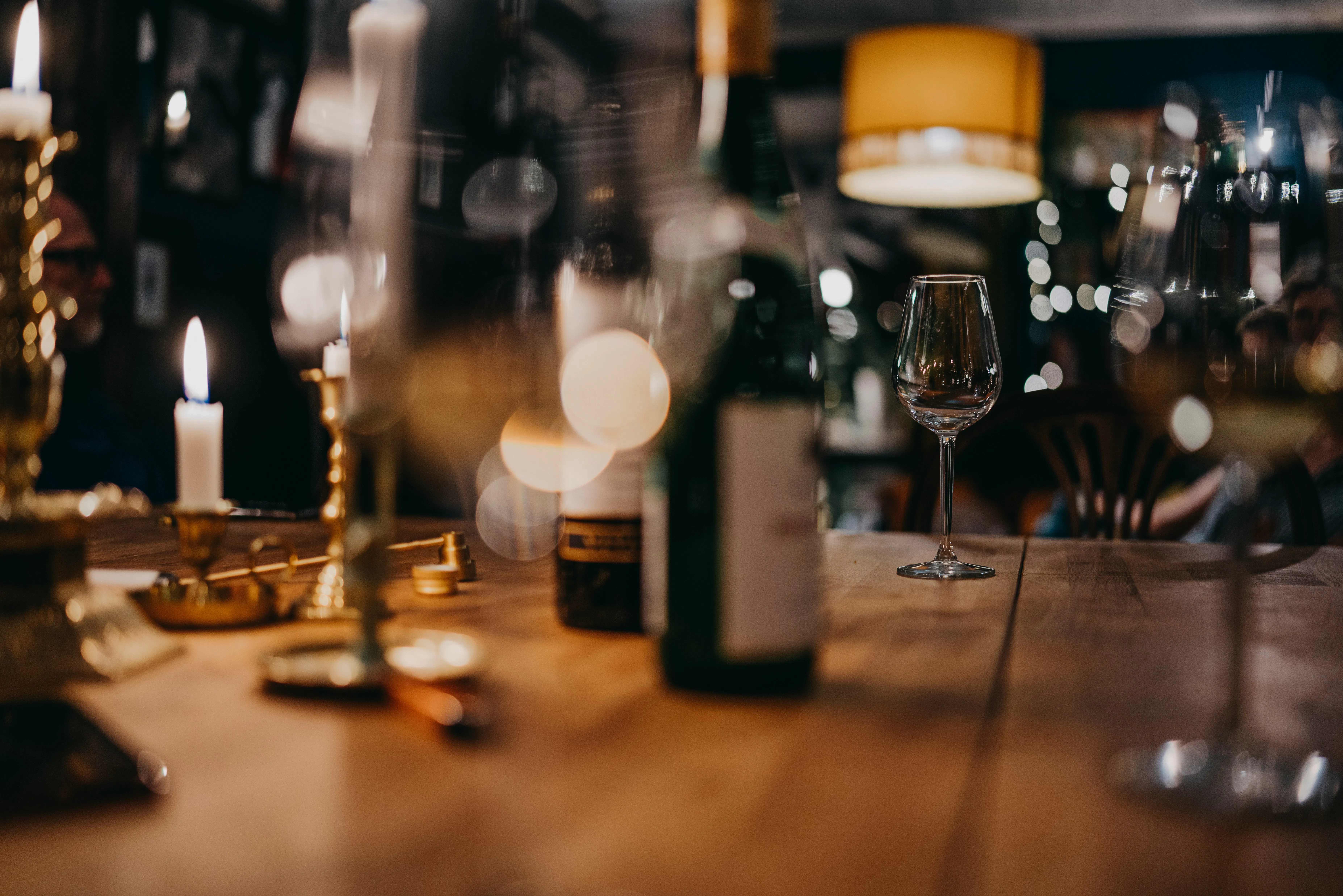 A moody, low-light shot of a rustic wooden dining table in a dimly lit room. The scene is warmly lit by glowing candles in brass holders and a yellow overhead lamp, with wine bottles and an empty wine glass in soft focus.