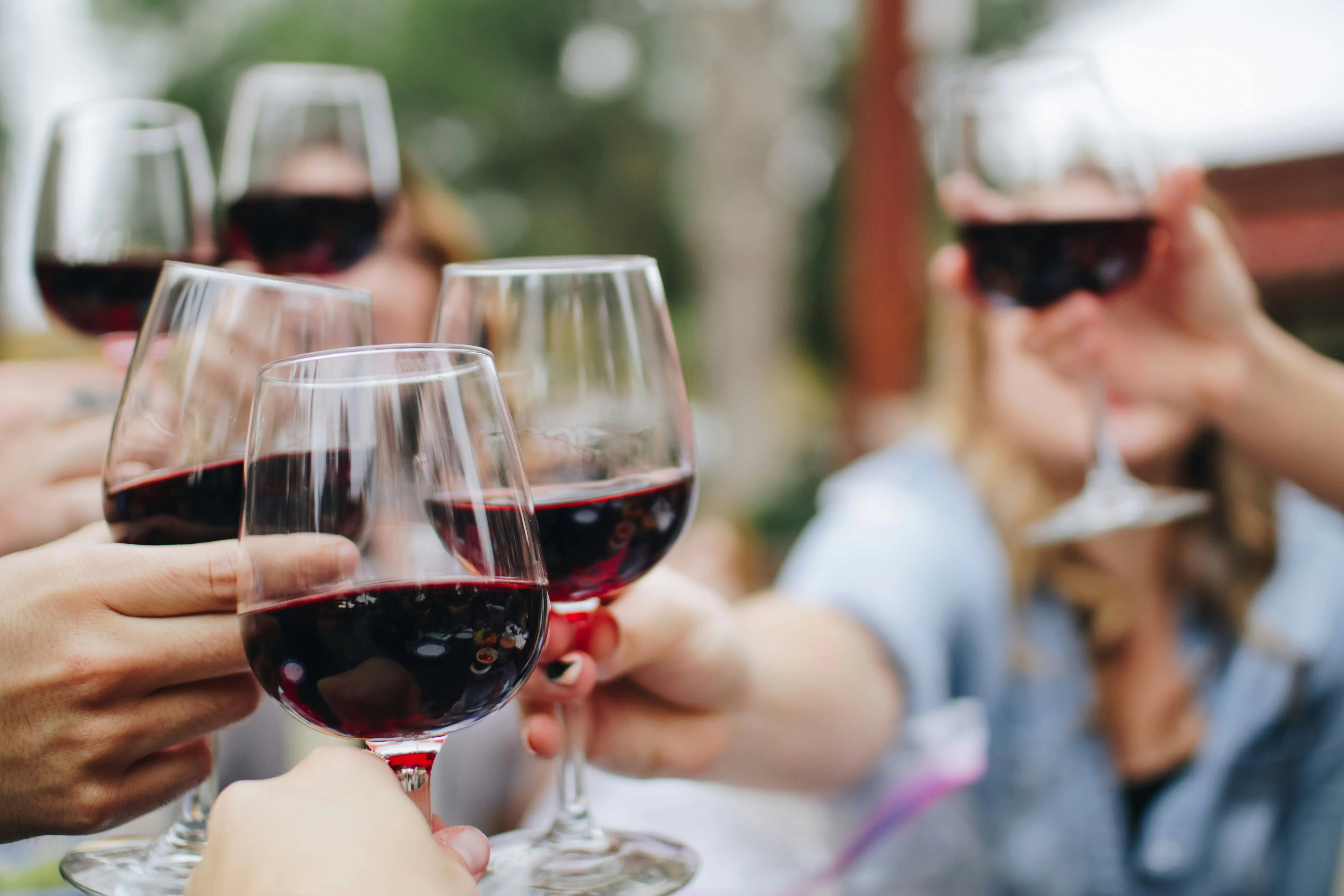 A close-up, slightly blurred shot of several people raising their glasses of red wine for a celebratory toast outdoors. The focus is on the rich, dark red wine inside the clear glasses.