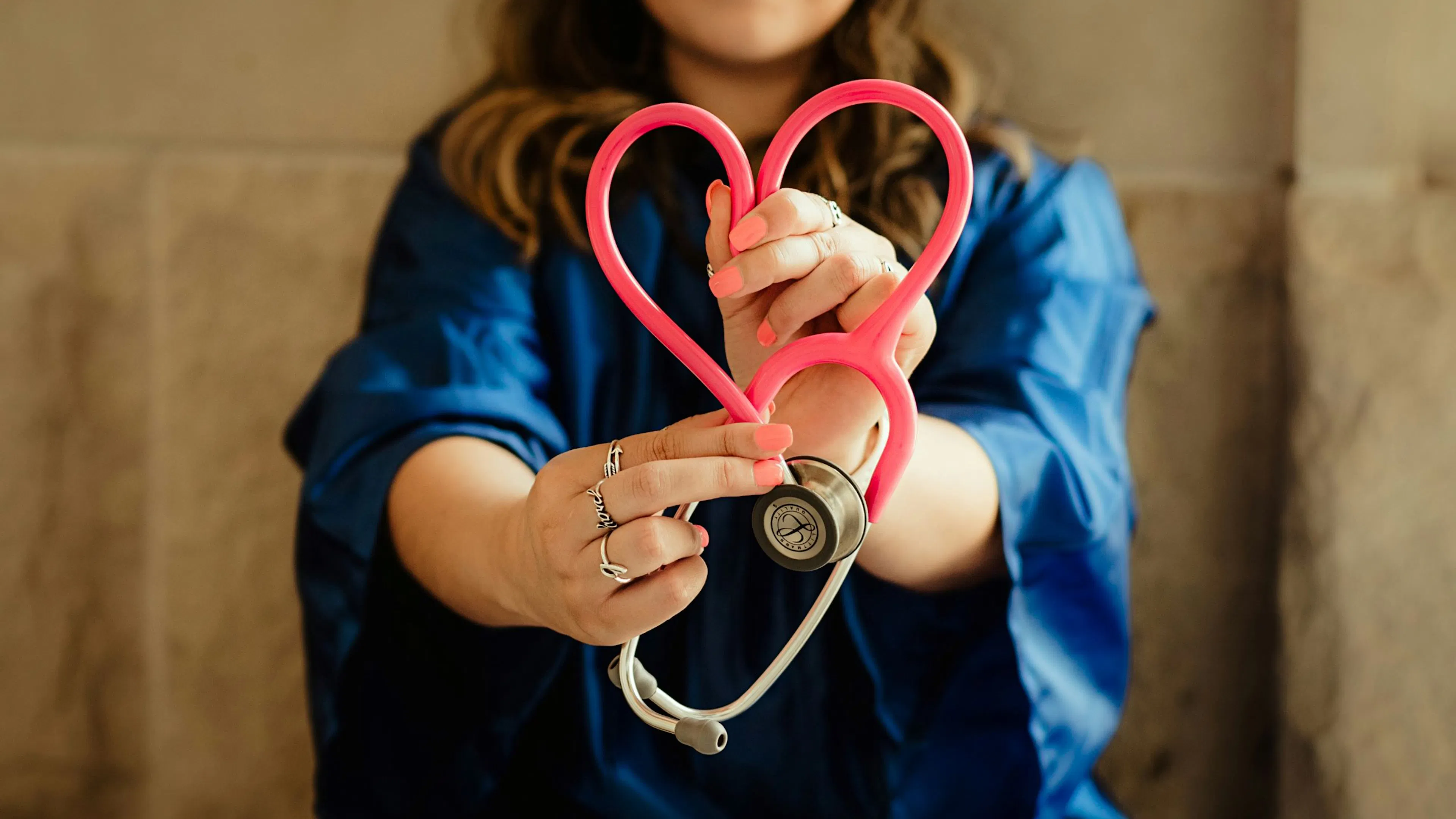 Woman in blue scrubs holding a pink stethoscope shaped like a heart