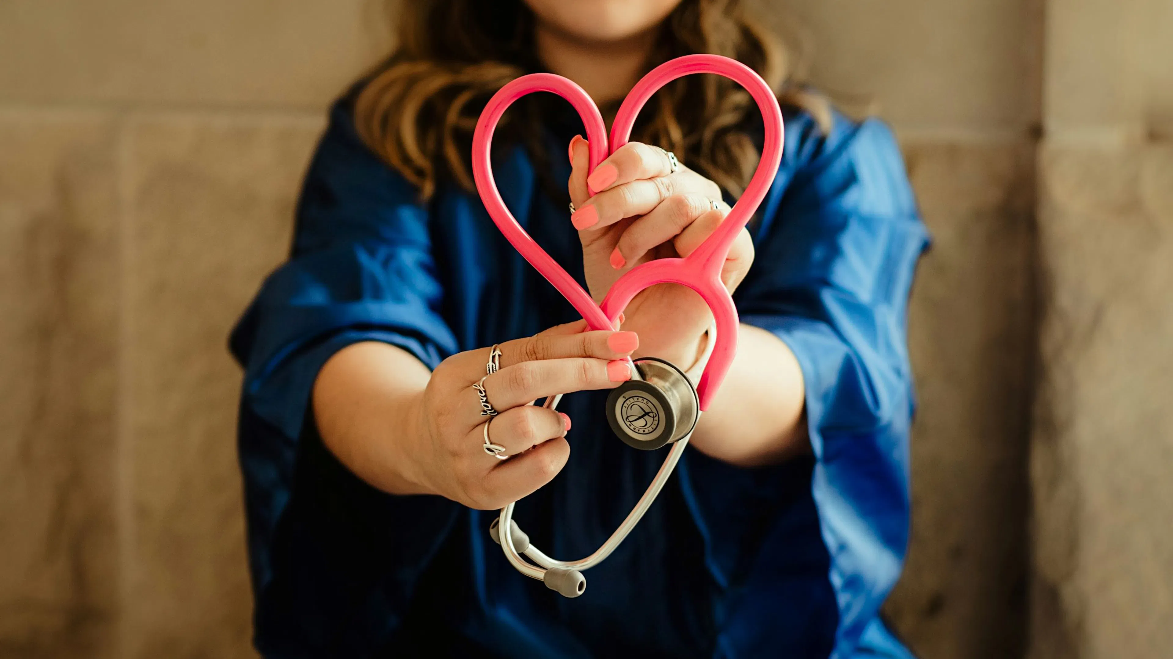 Woman in blue scrubs holding a pink stethoscope shaped like a heart