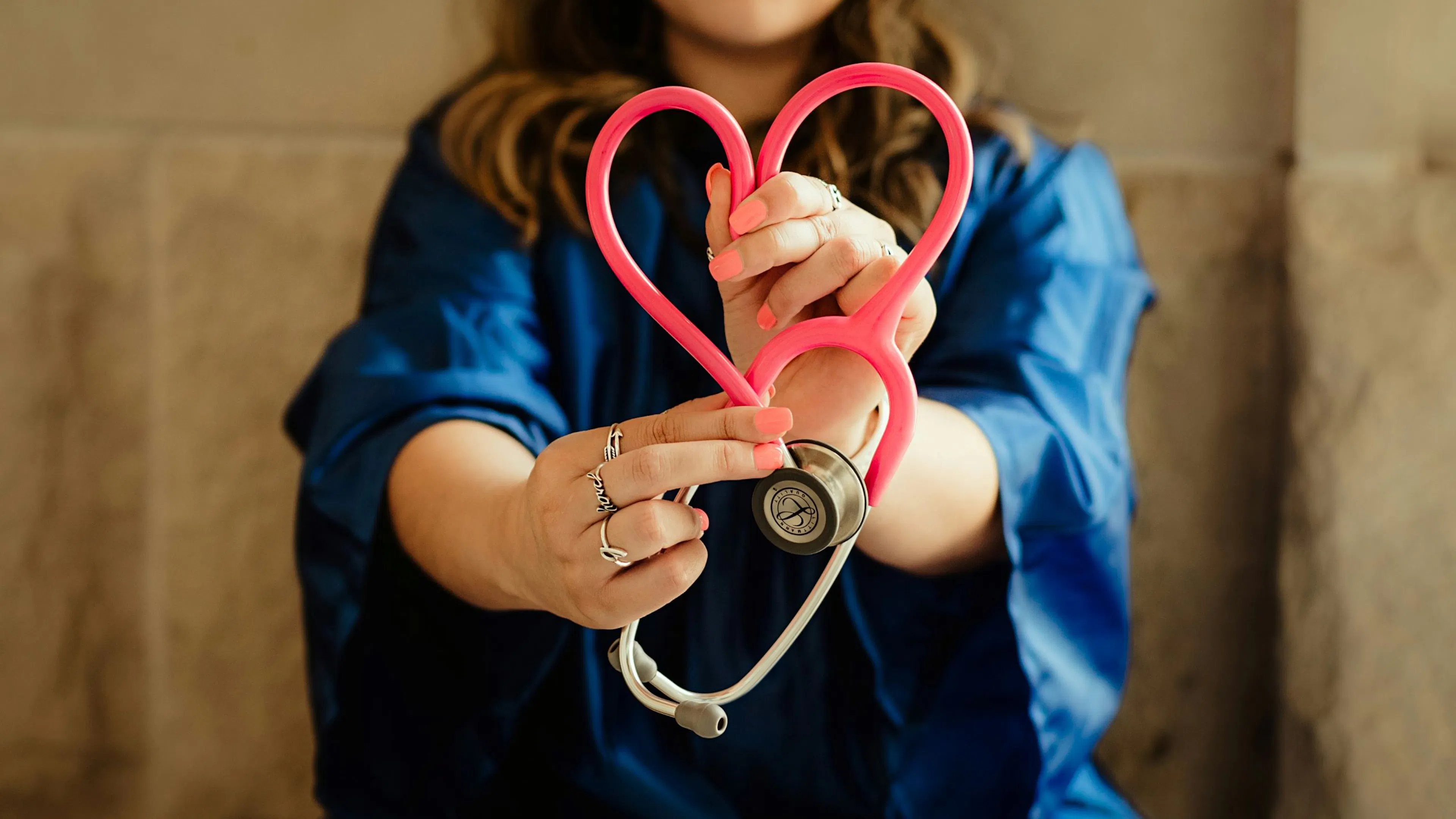 Woman in blue scrubs holding a pink stethoscope shaped like a heart