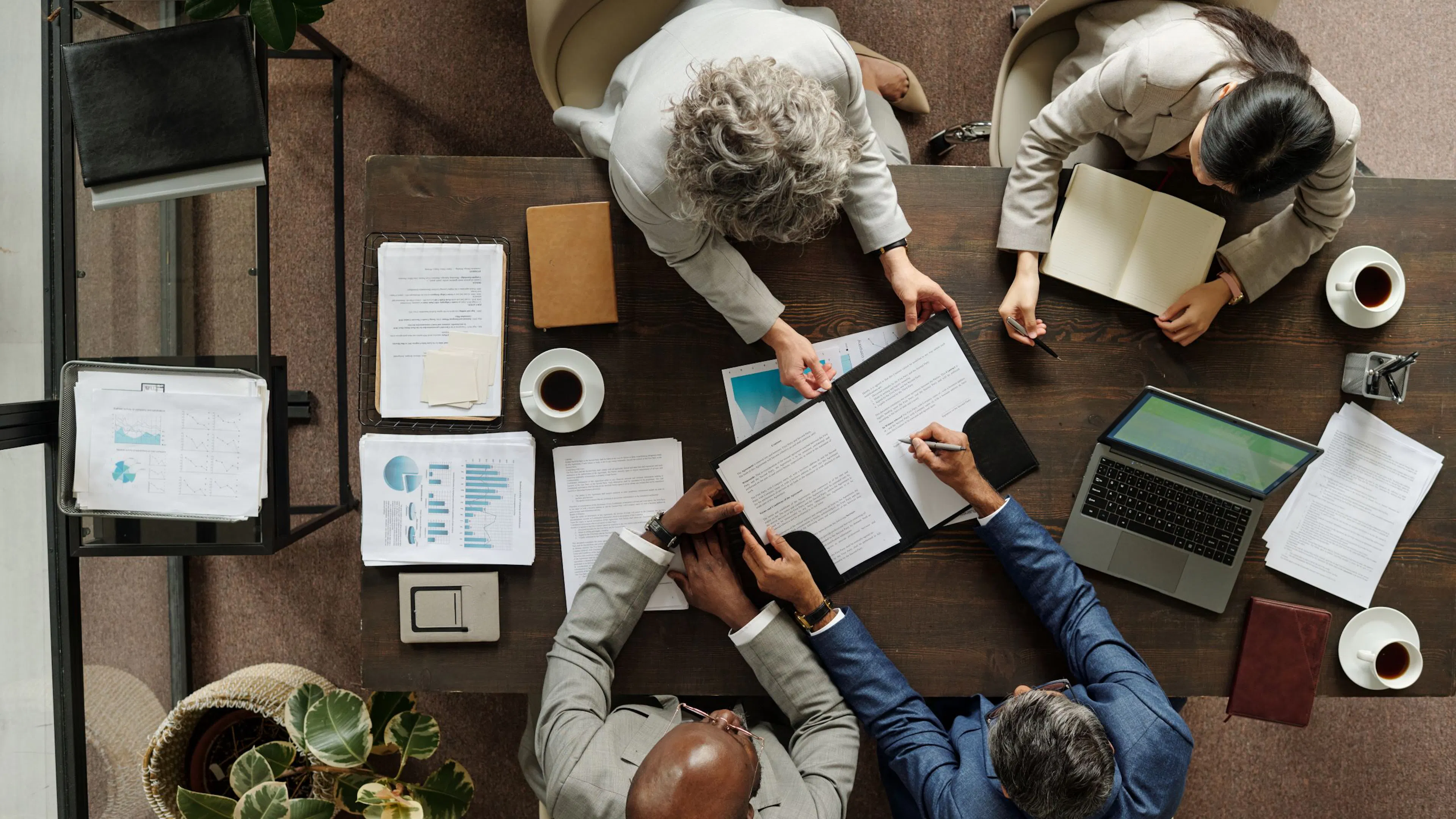 Overhead view of four professionals reviewing documents at a table with laptop, charts, and coffee during a meeting